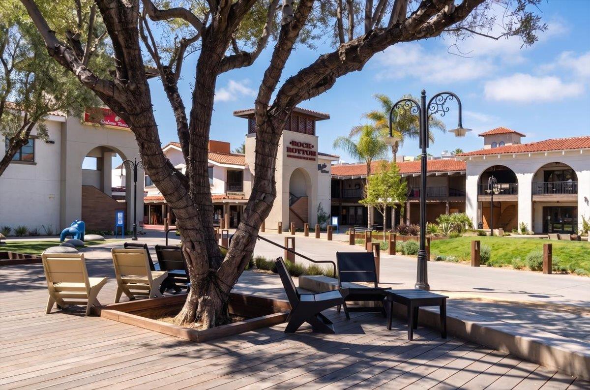 Shopping plaza with arched storefronts and outdoor seating beneath a large tree.