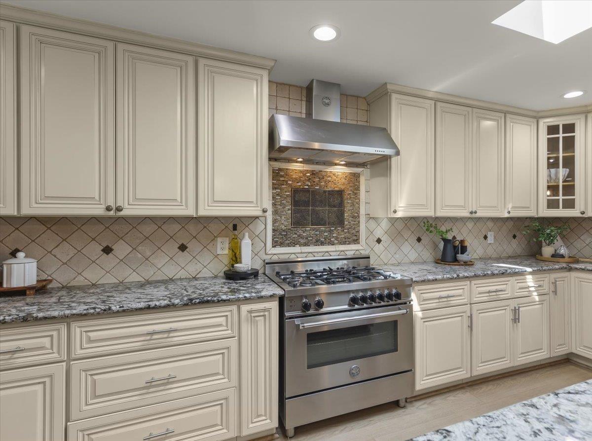 Kitchen with cream cabinetry, granite countertops, and a stainless-steel range beneath a broad metal hood.
