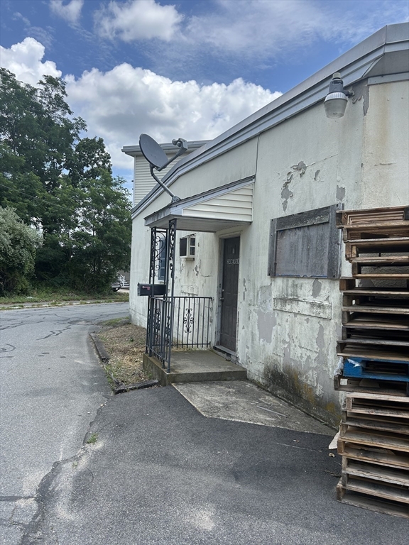 Weathered building exterior with a recessed entrance, metal railing, and stacked pallets beside the wall.