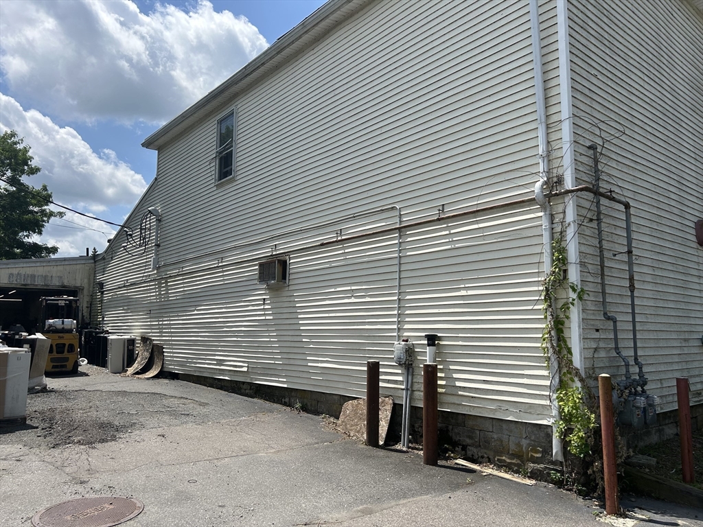 White-sided building with exposed utility pipes and meters along a paved side area.