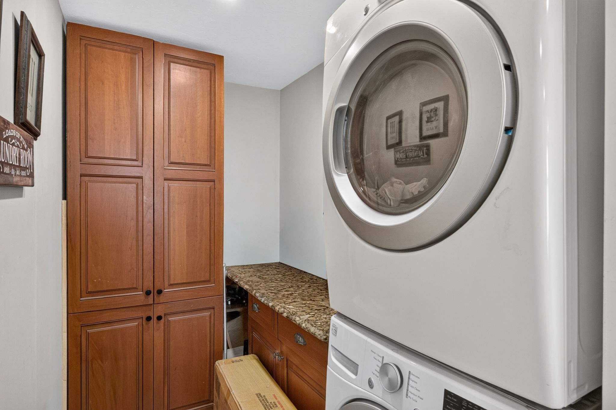 Laundry room with stacked front-loading washer and dryer beside wood cabinets and a granite counter.
