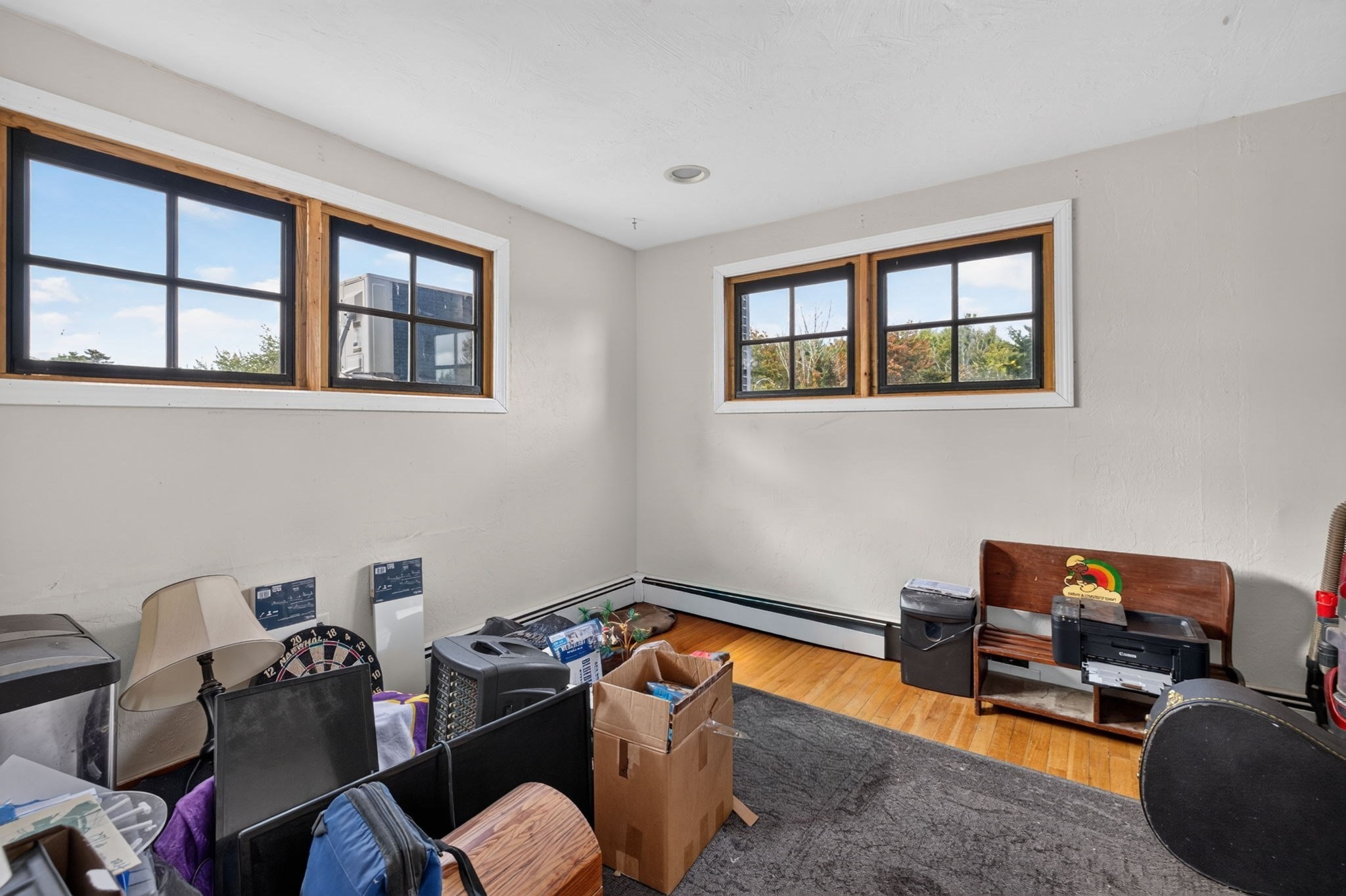 Cluttered room with hardwood flooring, gray carpet, boxes, and two sets of high windows.