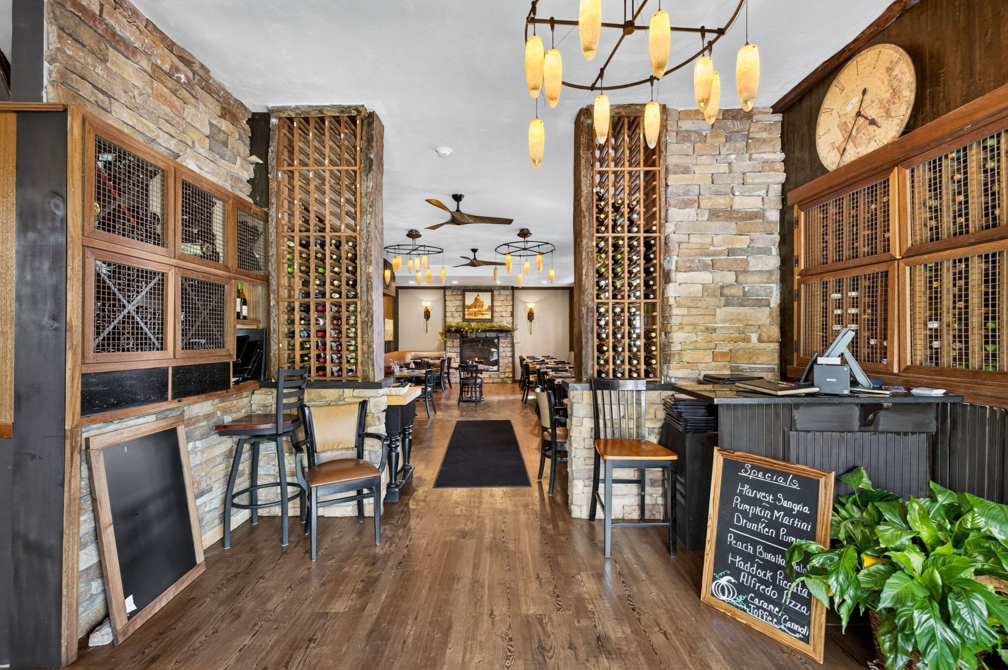 Restaurant dining room with stone walls, tall wine racks, pendant lights, and a wood service counter.