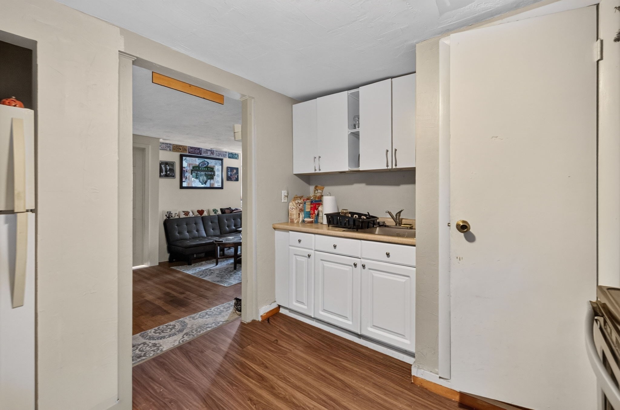 Compact kitchen with white cabinets and wood-look floors, opening onto a living room with a dark sofa.