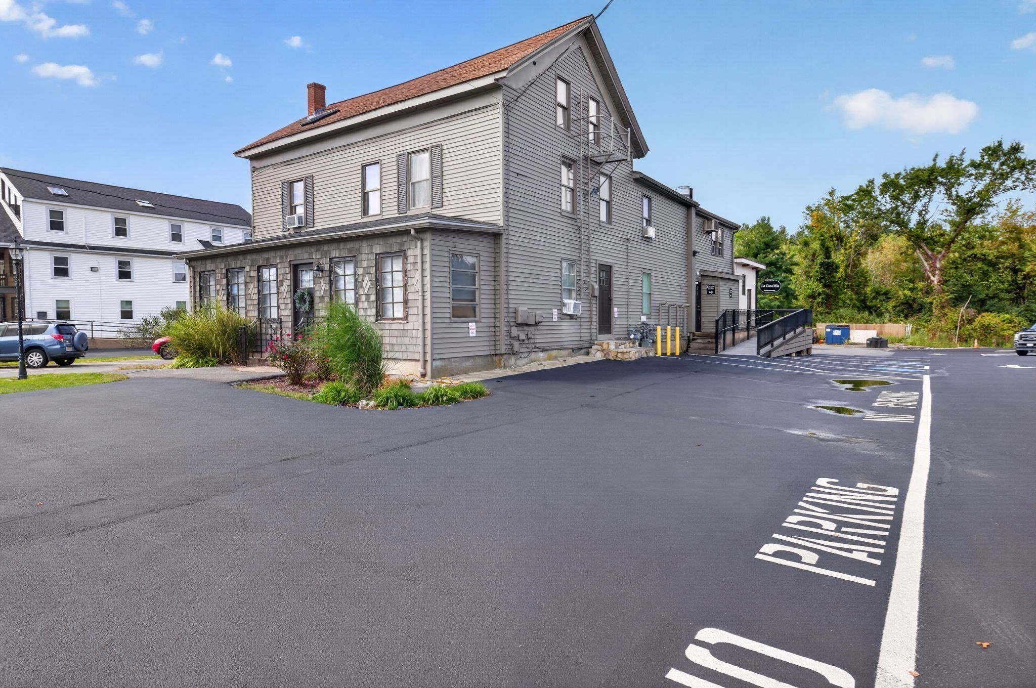 Gray multi-story building with a steep roof, upper balcony, accessibility ramp, and marked parking lot.