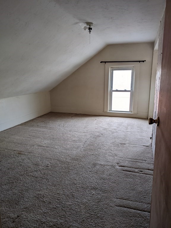 Empty attic bedroom with a sloped ceiling, gray carpet, and a single window.