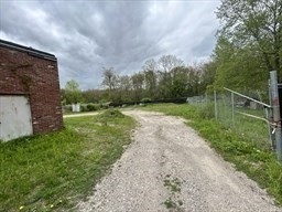 Gravel driveway through a grassy lot, with a brick building and chain-link fence.