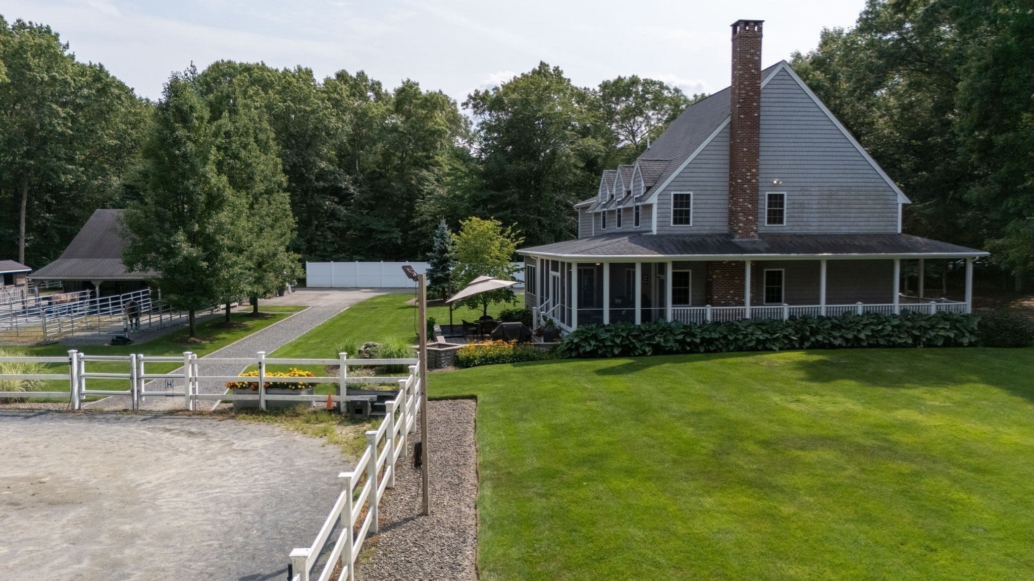 Gray shingle-sided house with a wraparound porch, lawn, white fencing, and a horse paddock.