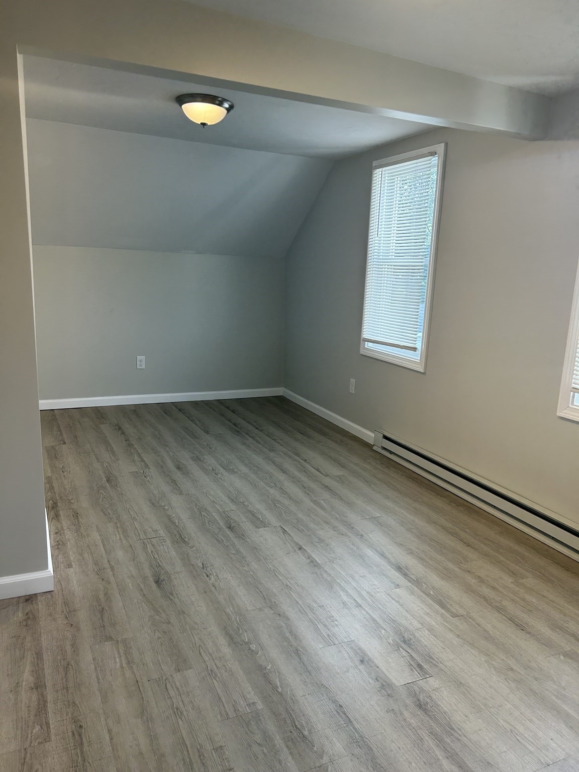 Empty room with gray wood-look flooring, a sloped ceiling, and a window with blinds.