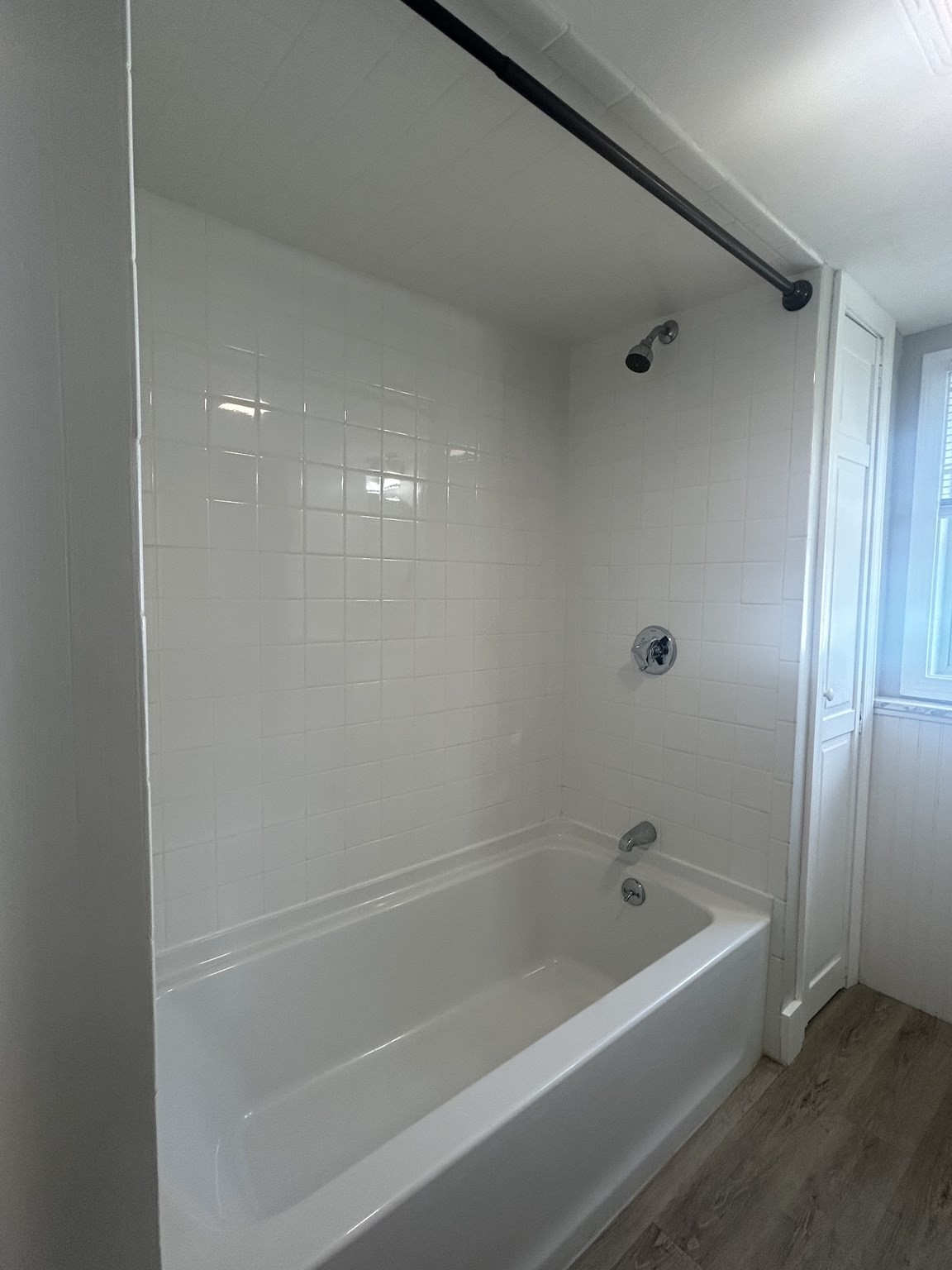 Bathroom with a white tub and shower, tiled surround, and wood-look flooring.