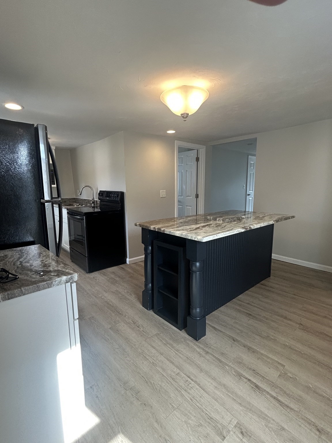 Kitchen with a large dark island, patterned stone counters, black appliances, and wood-look flooring.