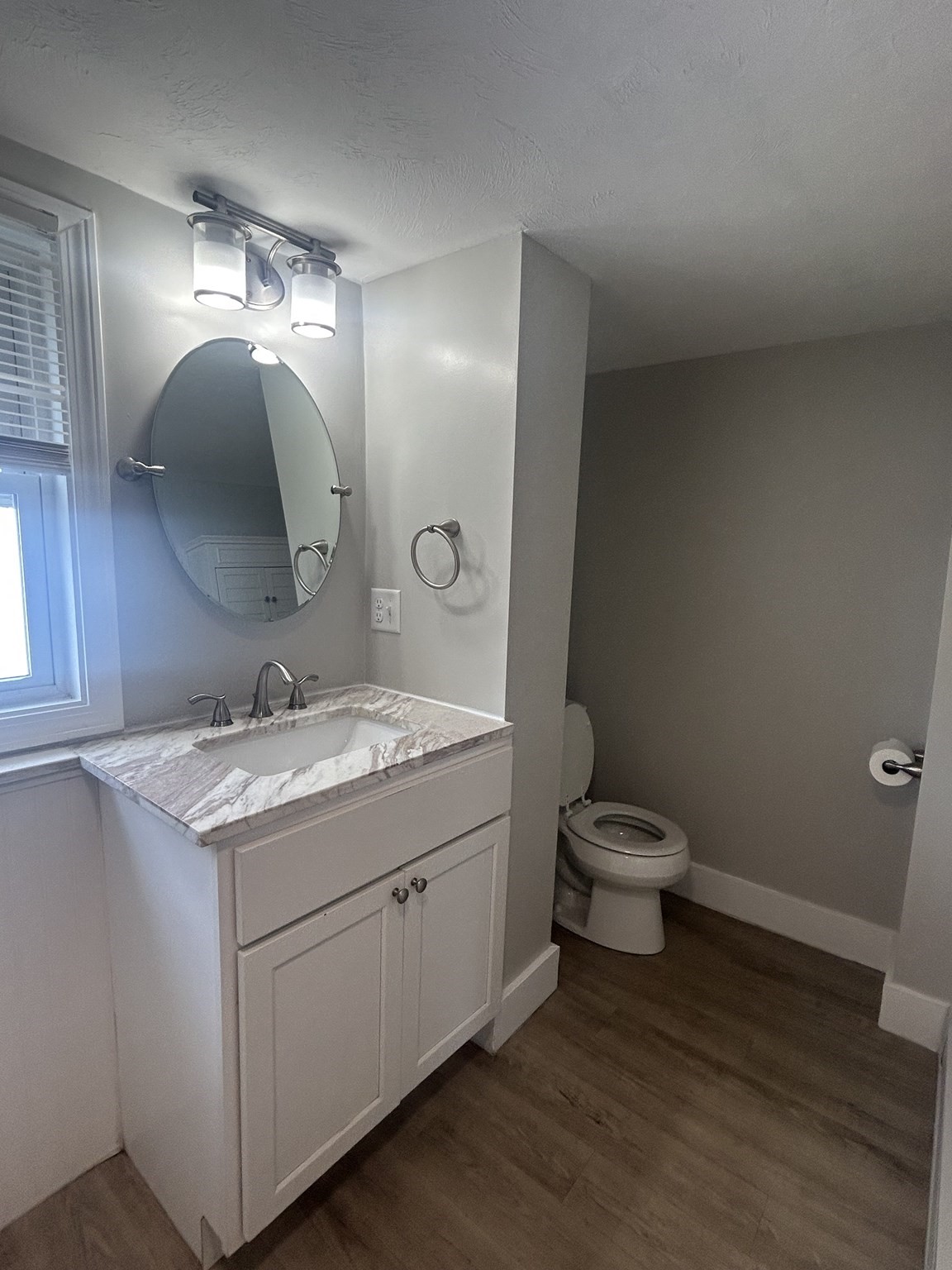 Bathroom with a marble-look vanity, oval mirror, window, and toilet alcove.