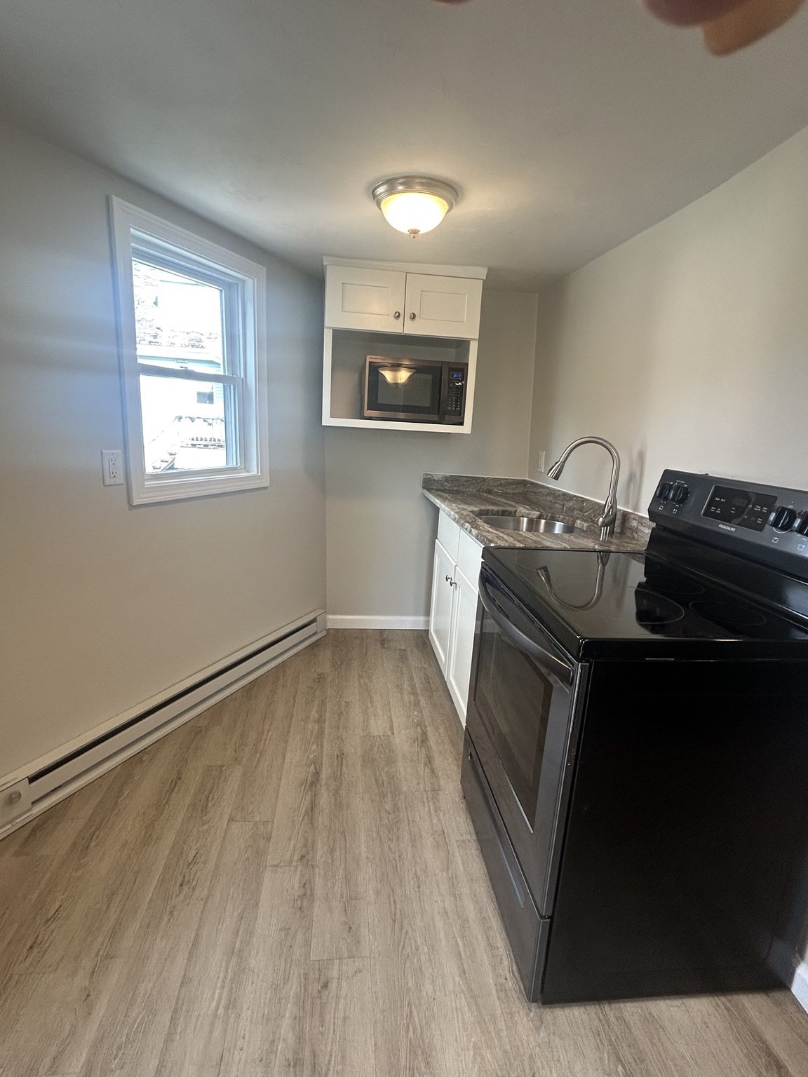 Narrow kitchen with white cabinets, black electric range, stainless sink, and wood-look flooring.