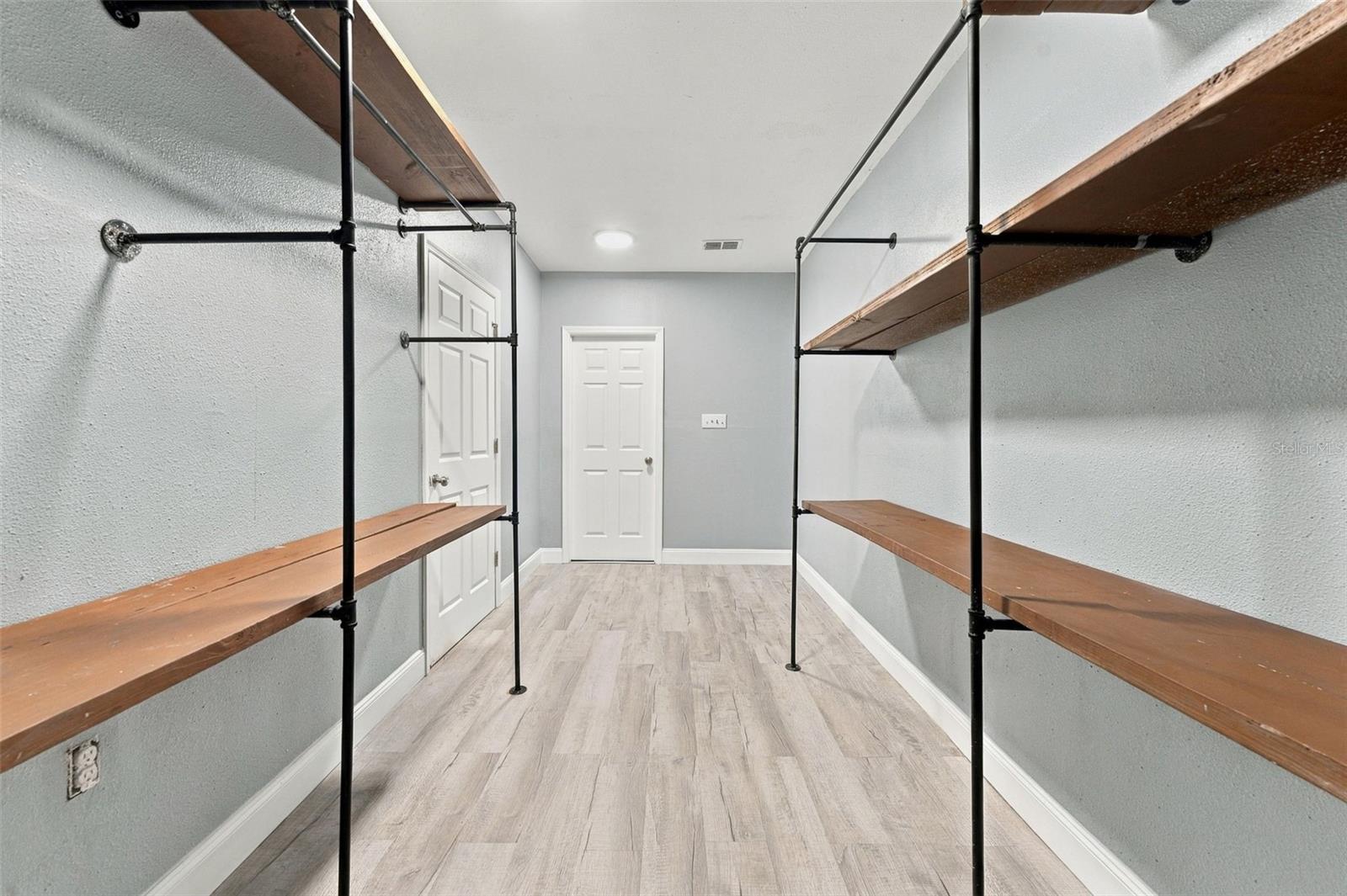 Walk-in closet with open wooden shelving on both sides and light wood-look flooring.