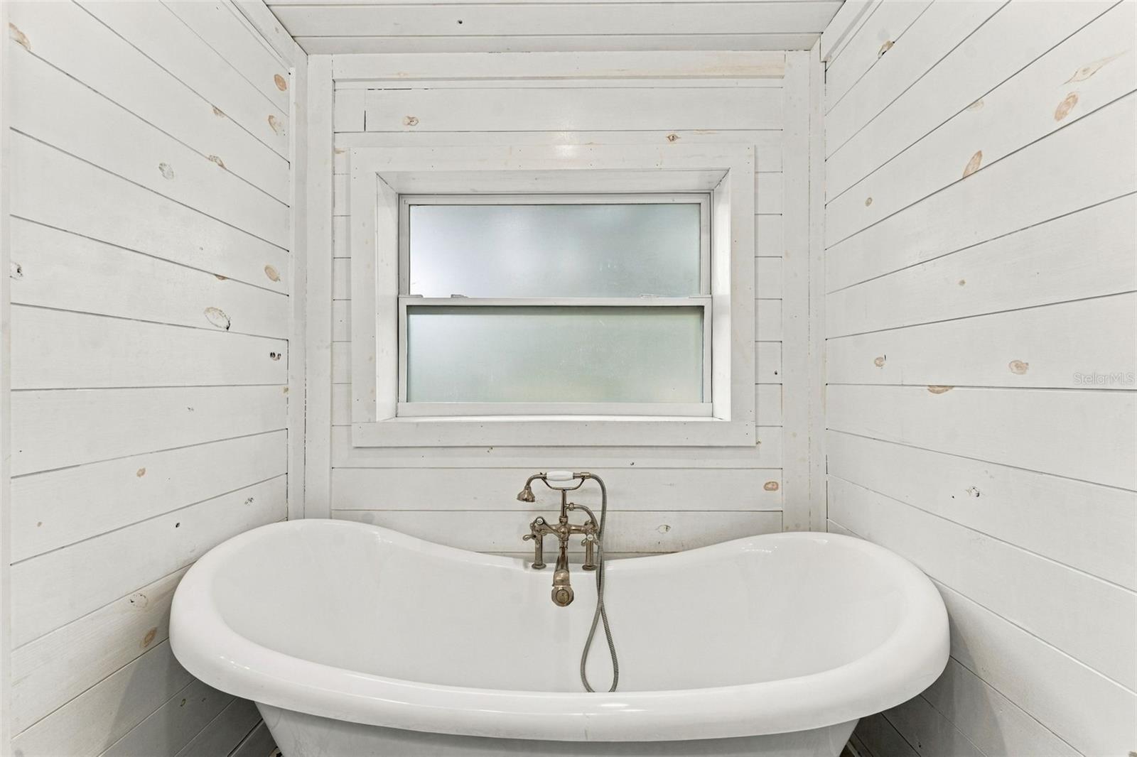 White freestanding bathtub with antique-style brass faucet beneath a frosted window, surrounded by white wood paneling.