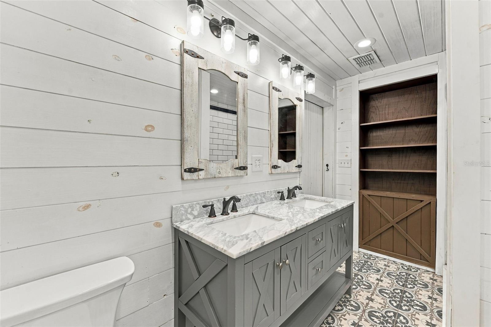 Bathroom with a double-sink gray vanity, rustic mirrors, patterned tile floor, and built-in wooden shelving.