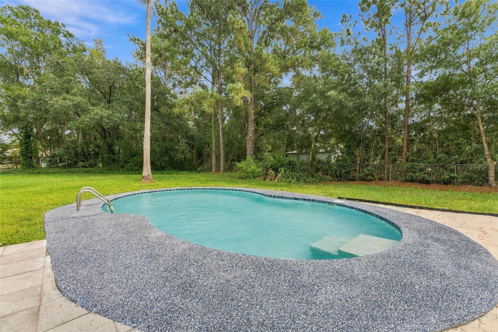 Backyard swimming pool with a curved blue-speckled deck, surrounded by lawn and mature trees.