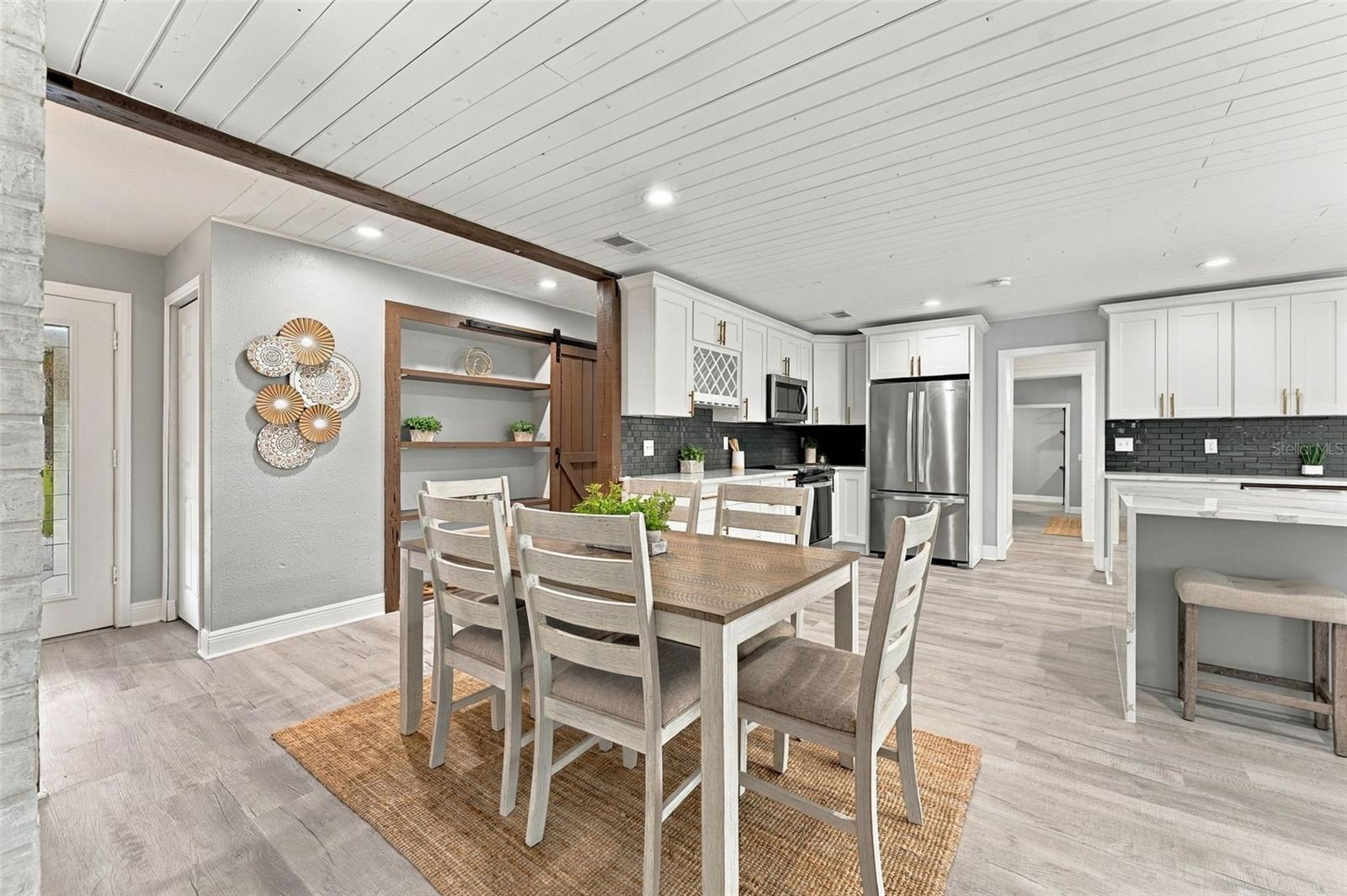 Dining area and kitchen with white cabinets, dark backsplash, stainless appliances, and a wood-beam ceiling.