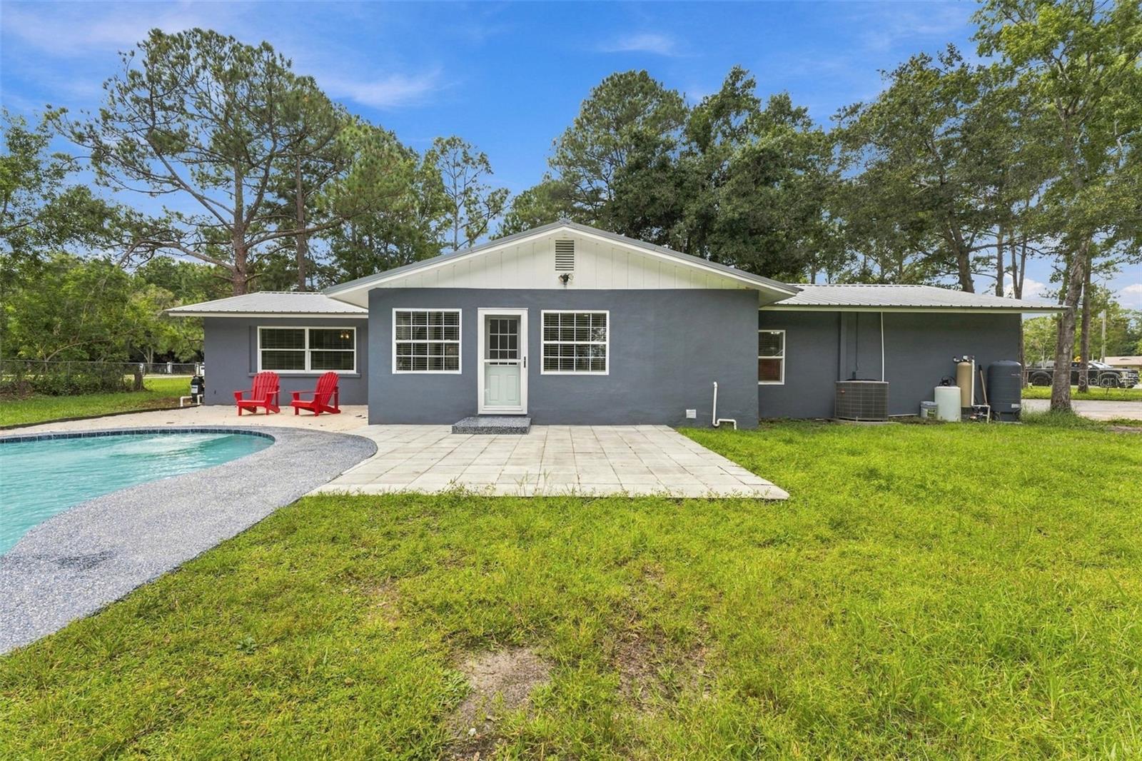 Rear of a gray single-story home with a curved swimming pool and red patio chairs.