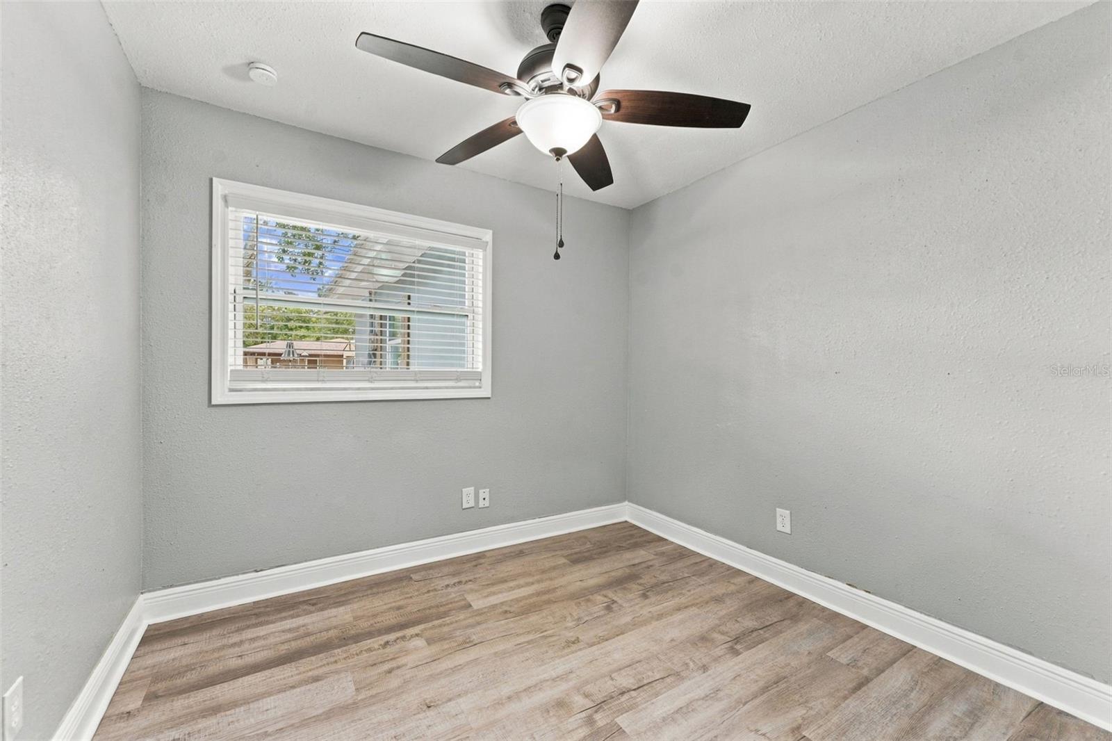 Empty room with gray walls, wood-look flooring, and a ceiling fan beneath a window with blinds.