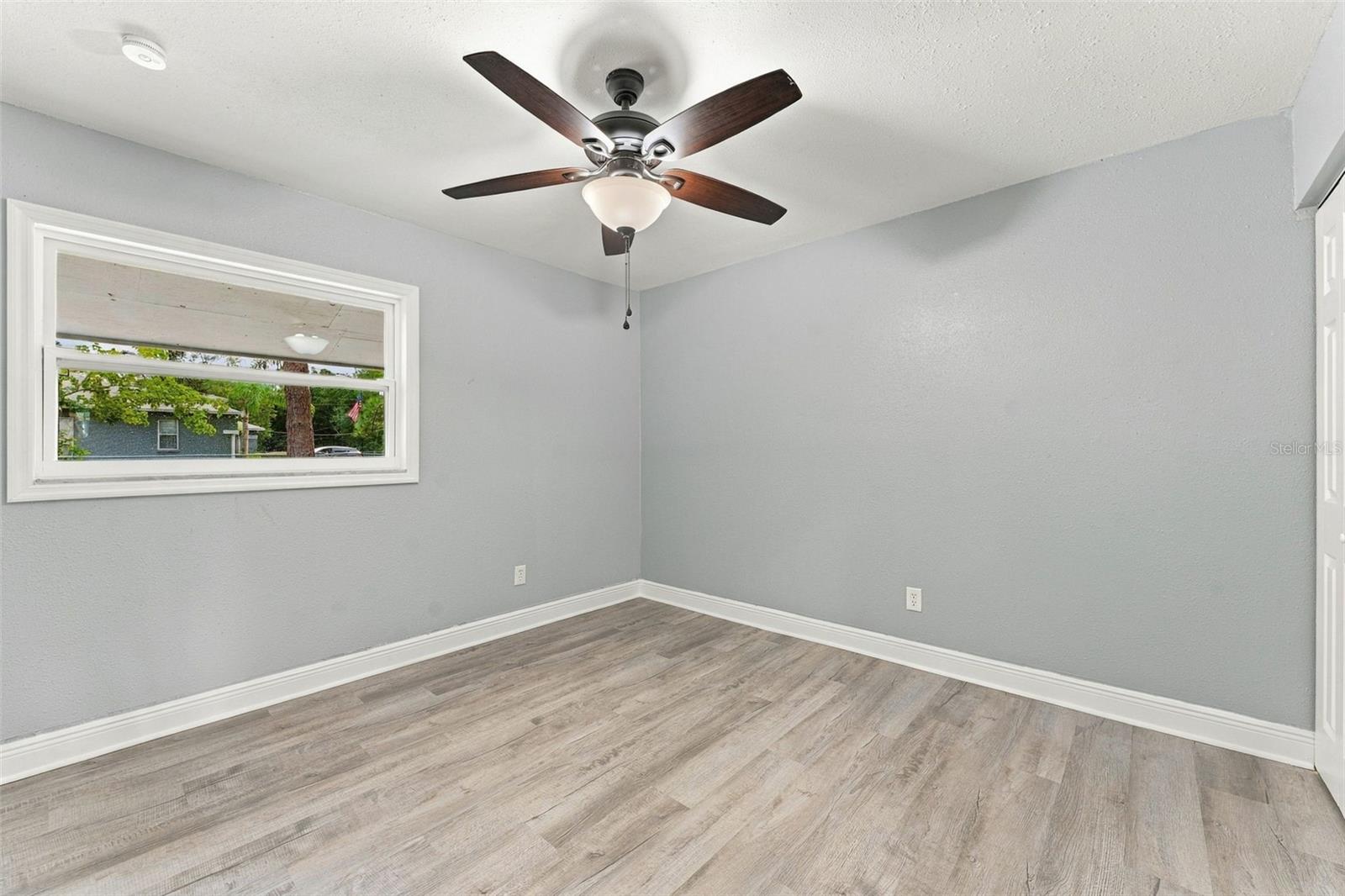 Empty room with gray plank flooring, a wide window, and a dark ceiling fan with a light.