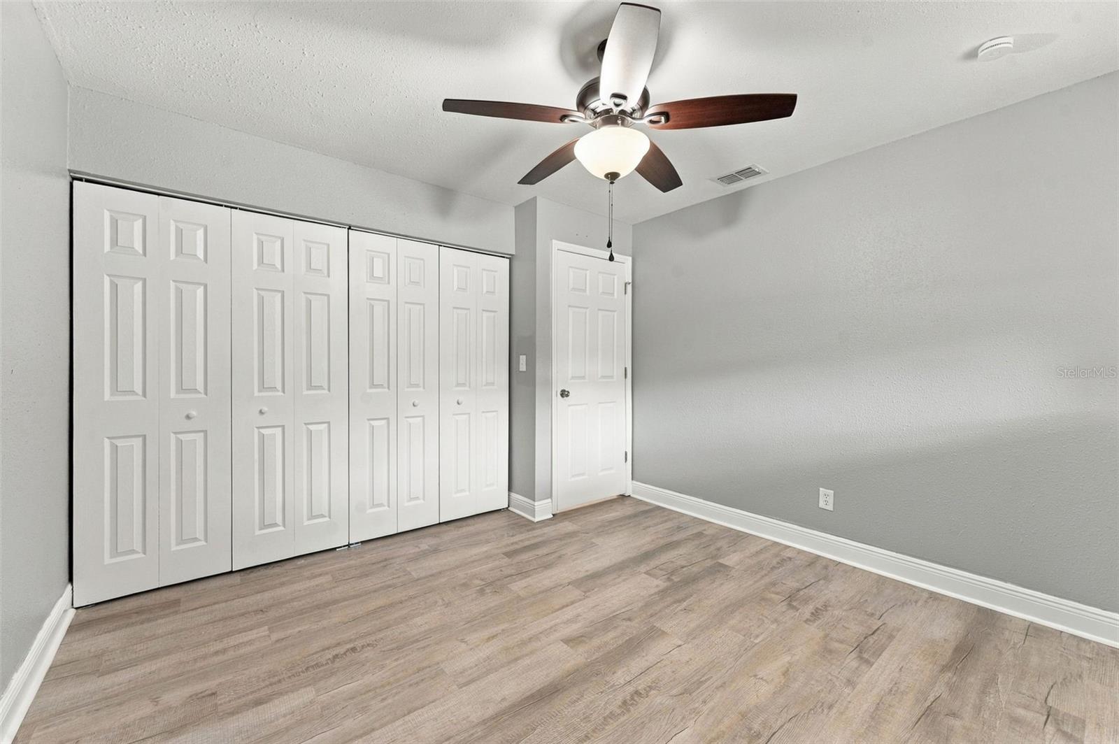 Empty bedroom with white bifold closet doors, wood-look flooring, and a ceiling fan with light.
