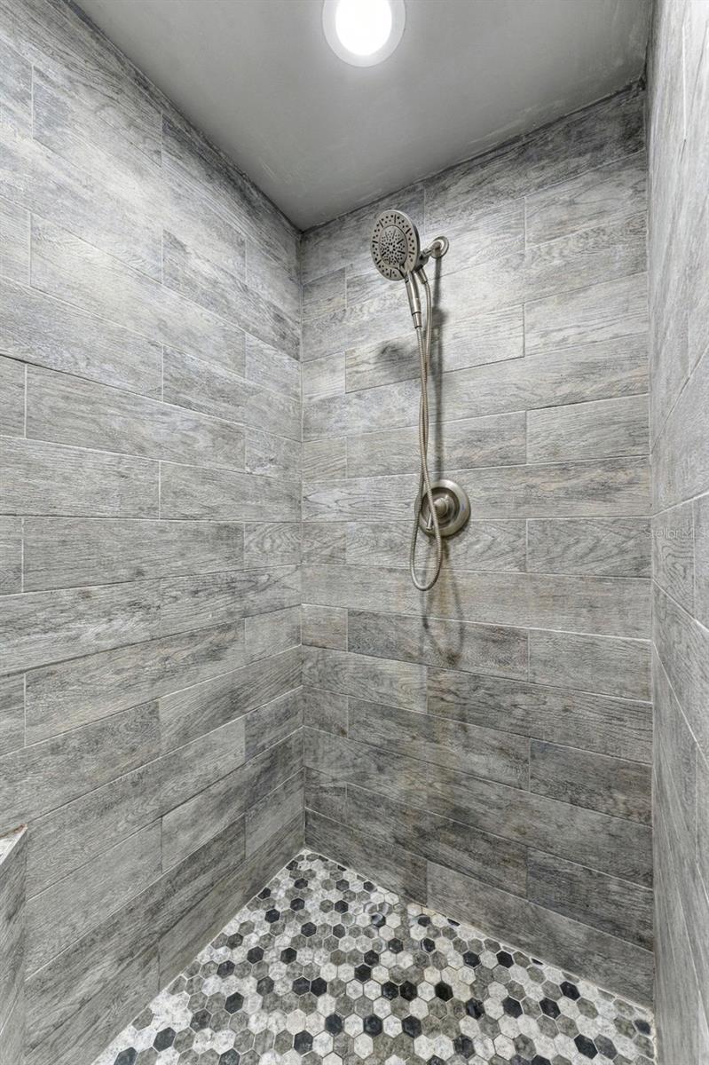 Walk-in shower with gray wood-look tile, handheld showerhead, and black-and-white hexagonal mosaic floor.