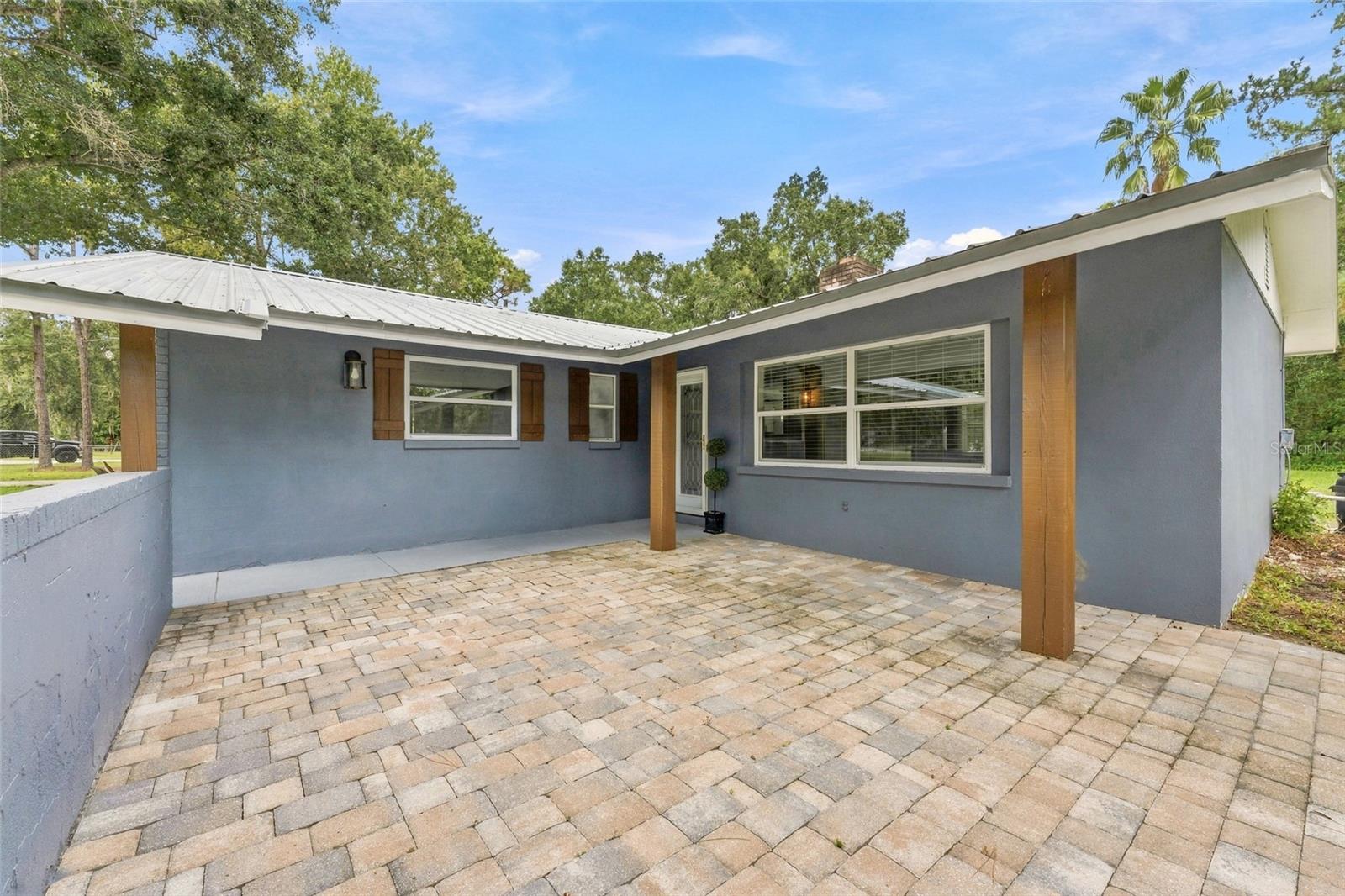 Gray stucco home's covered entry patio with brick pavers and a white metal roof.