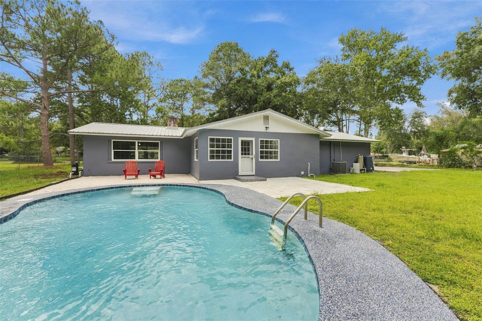 Single-story gray home behind a curved swimming pool, with a patio, two red chairs, and mature trees.