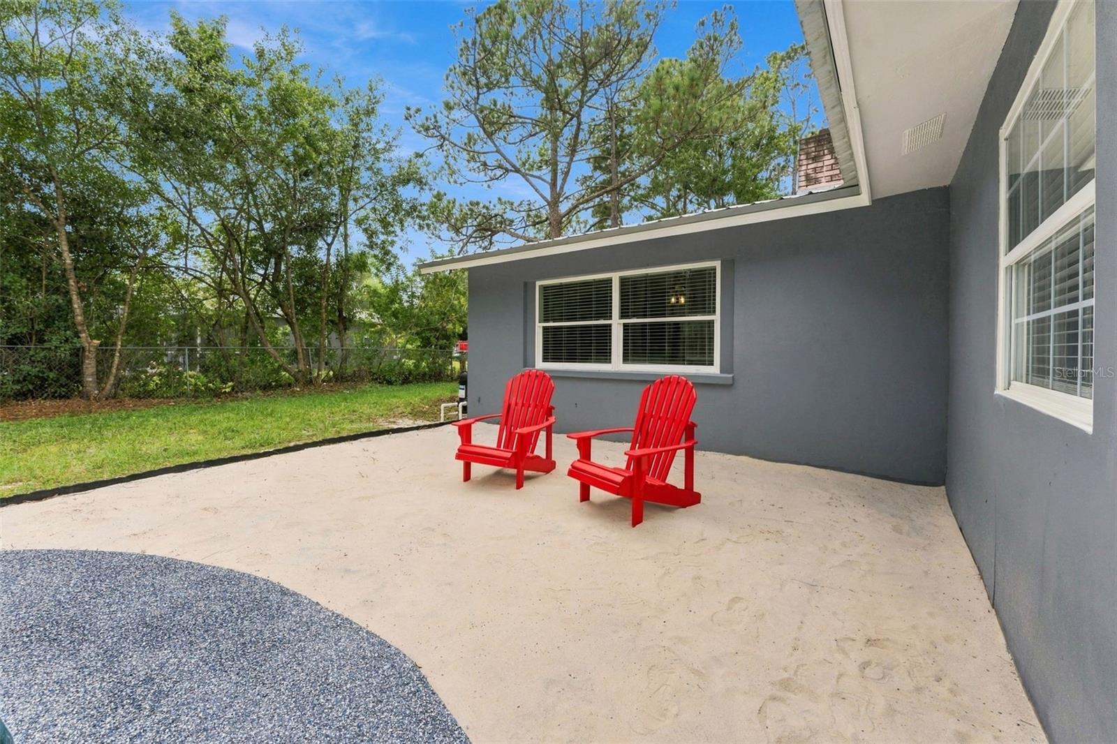 Concrete patio beside a gray home, with two red Adirondack chairs and trees along the yard.