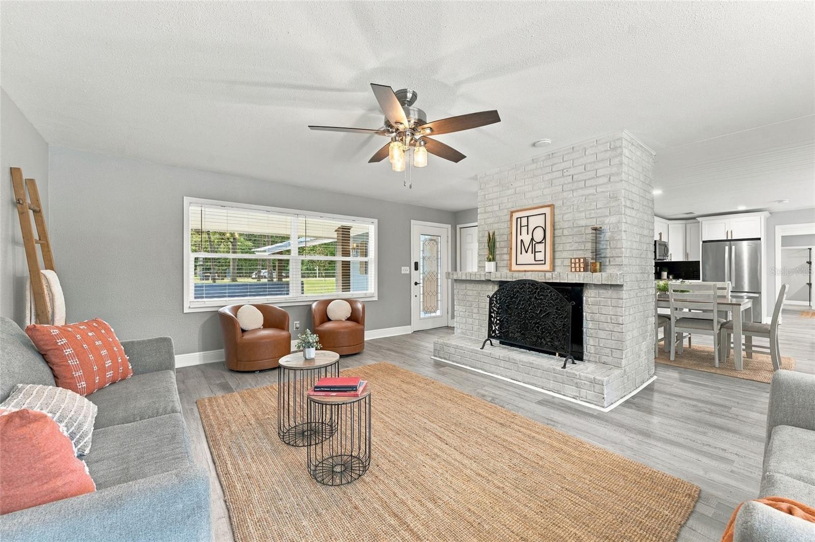 Staged living room with a white brick fireplace, gray seating, and an adjoining dining area.