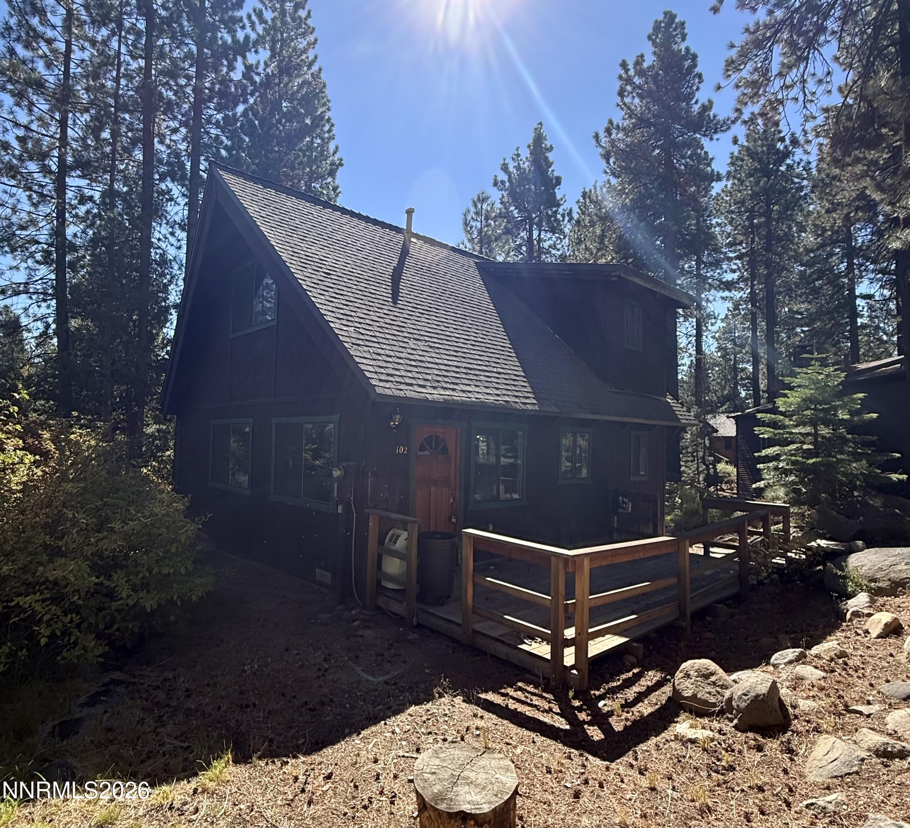 Dark A-frame cabin with a small wooden deck among tall pines and rocks.