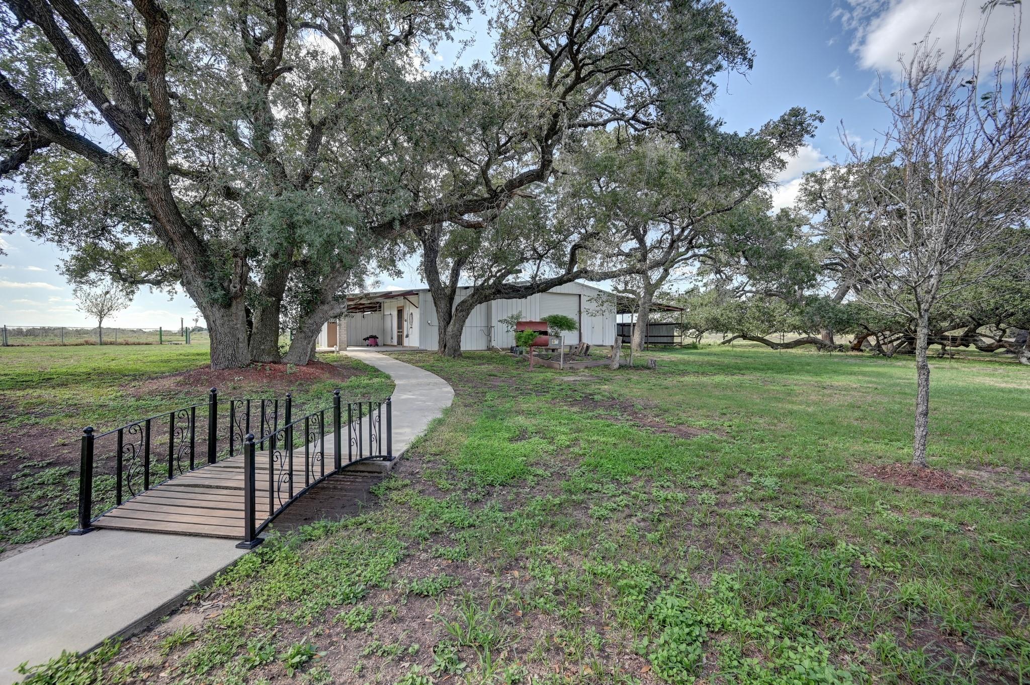 Single-story house beneath mature oak trees, approached by a curved walkway and small black-railed bridge.
