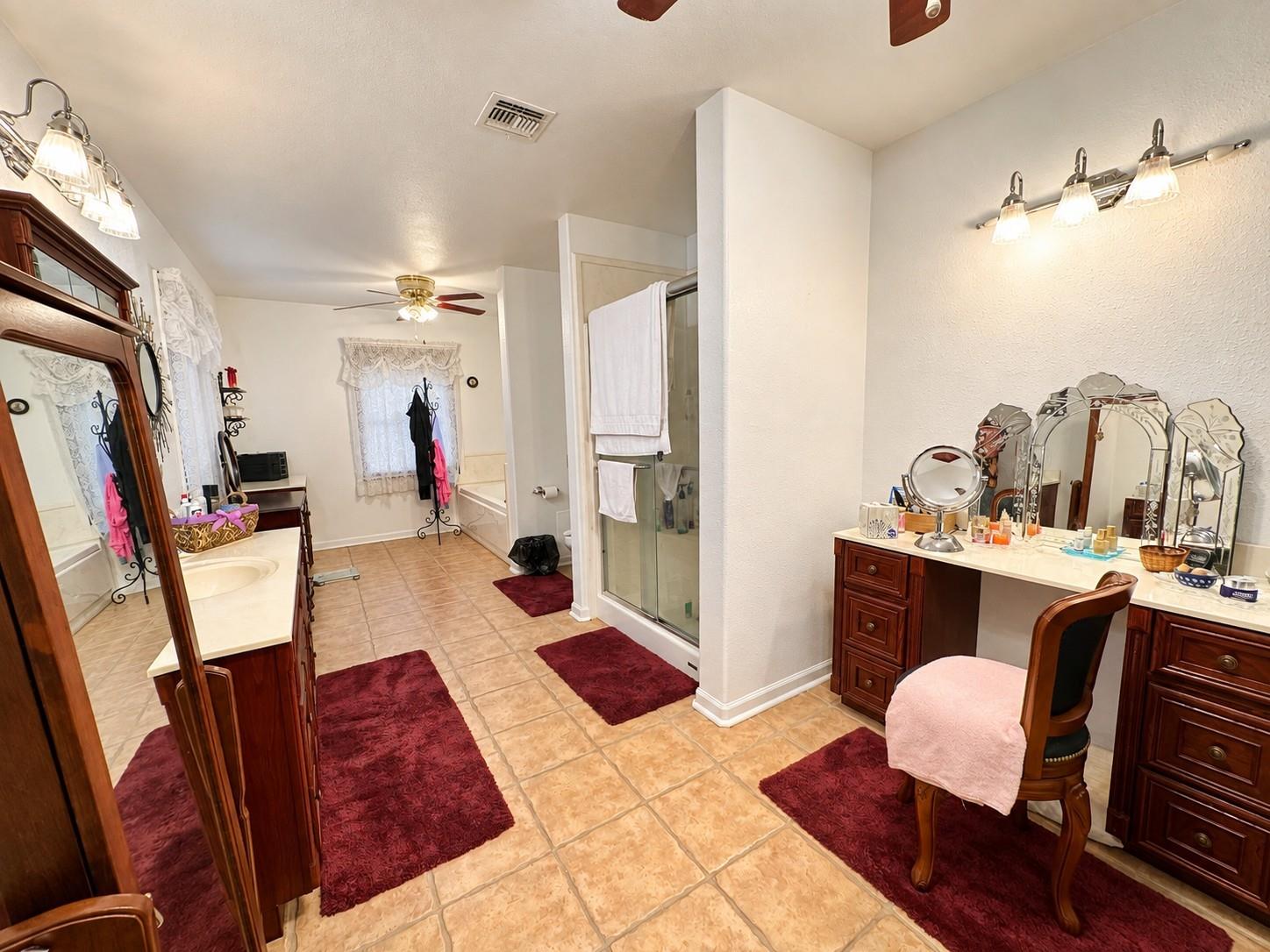 Spacious bathroom with dual wood vanities, a glass shower, bathtub, and burgundy rugs.