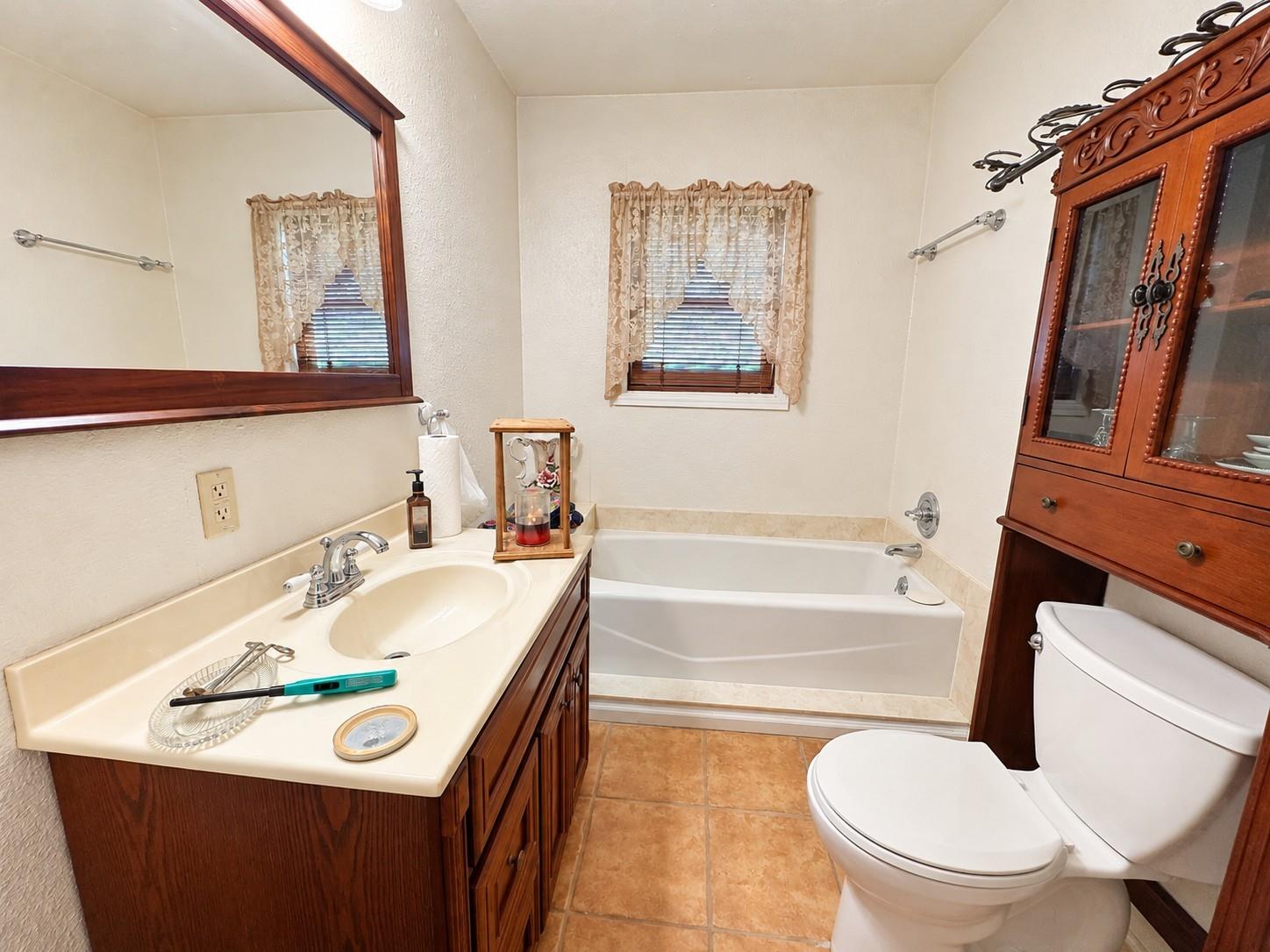 Bathroom with a tub beneath a curtained window, wood vanity, and carved wooden cabinet.