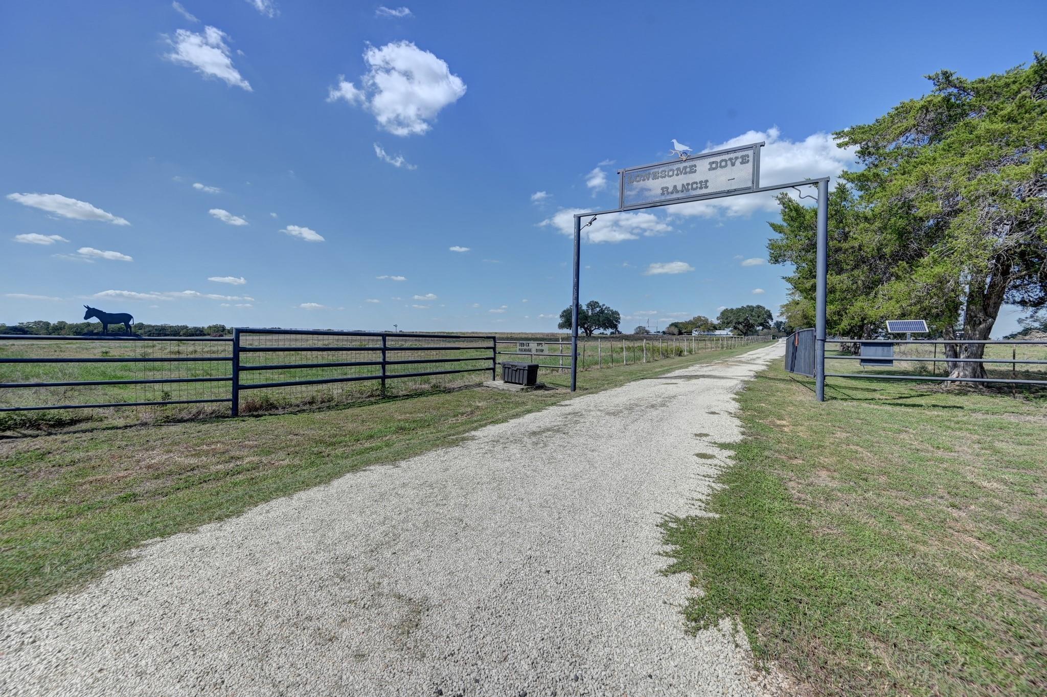 Ranch entrance with a gravel drive, metal Lonesome Dove Ranch sign, fenced pasture, and donkey silhouette.