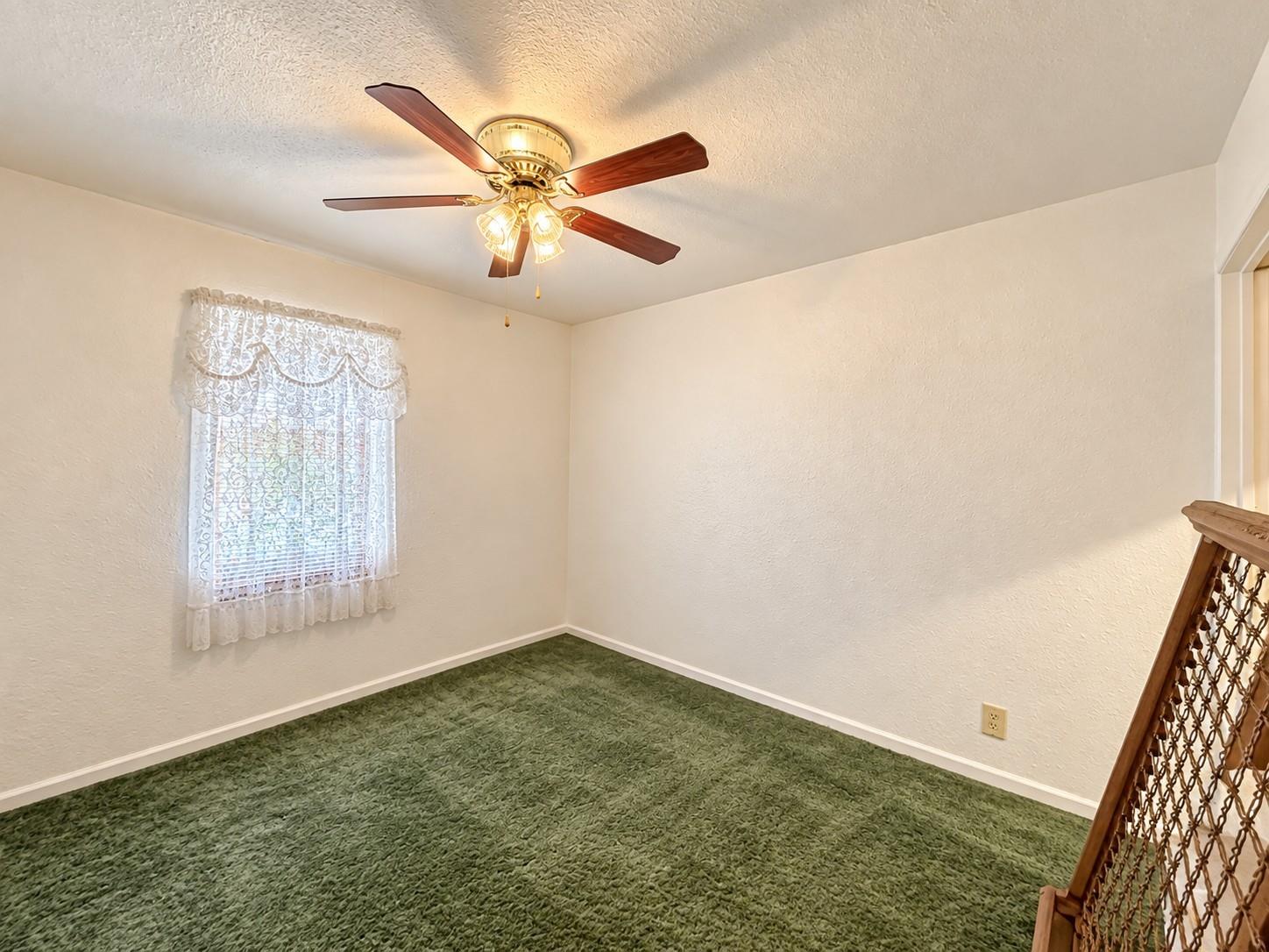 Empty bedroom with green carpet, a curtained window, and a lit ceiling fan.