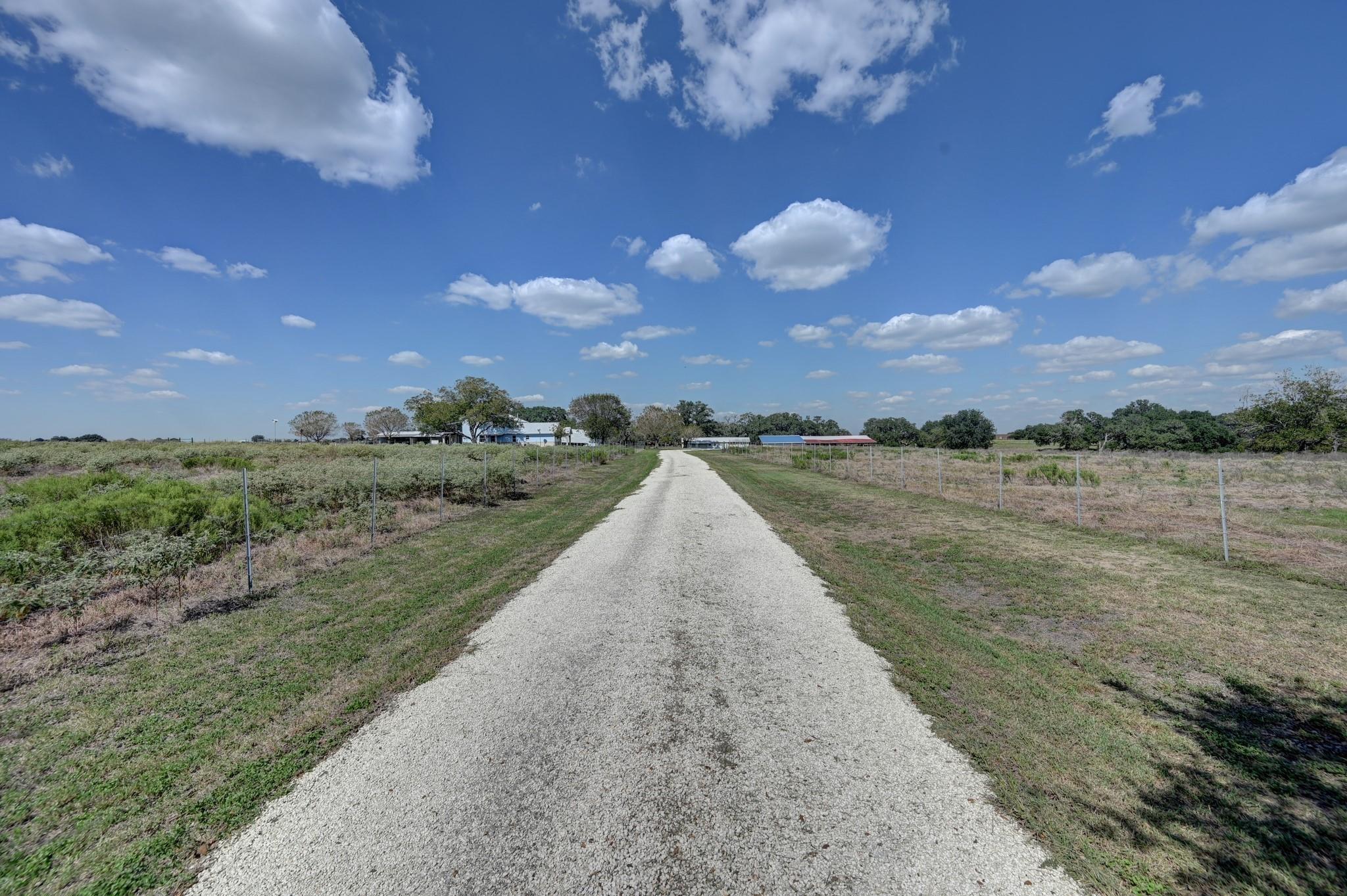 Gravel driveway crosses fenced fields toward rural buildings and scattered trees.