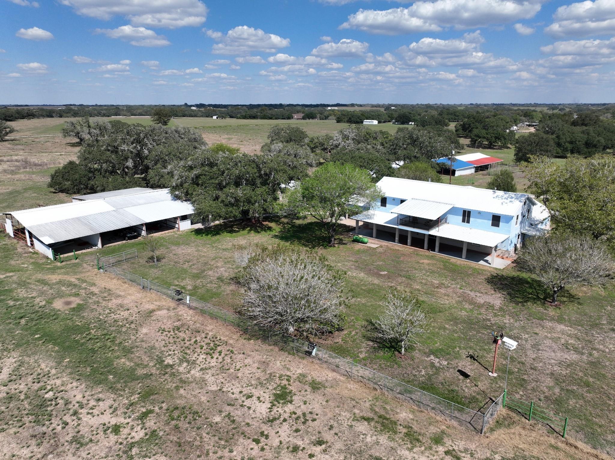 Aerial view of a light-blue raised house and large metal-roof barn surrounded by fenced pasture and mature trees.