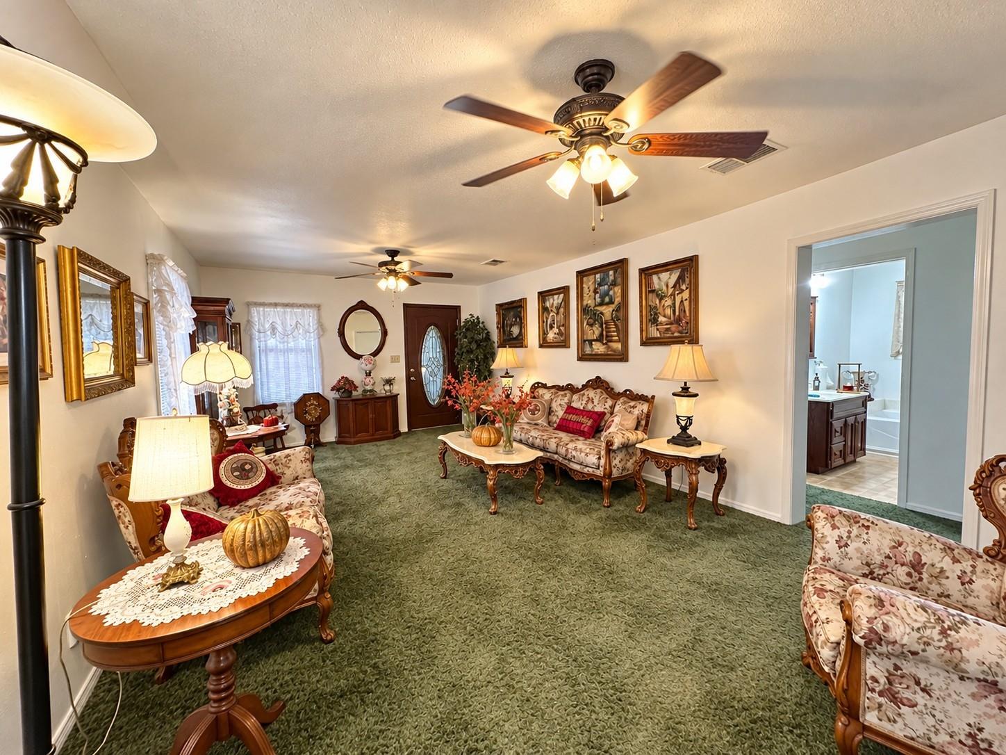 Furnished living room with green carpet, ornate seating, carved wood tables, and a doorway to the bathroom.