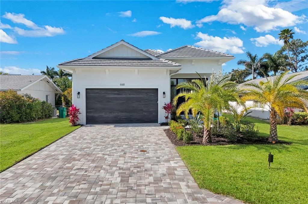Modern white home with a dark double garage, paver driveway, and tropical landscaping.