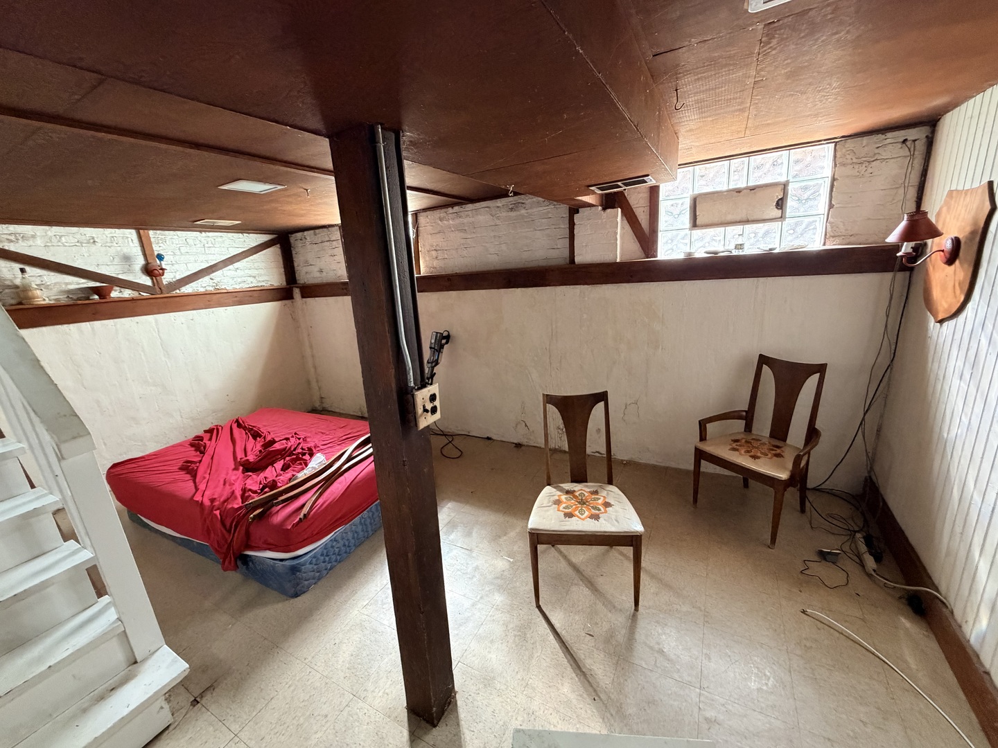Basement bedroom with red bedding, exposed beams, two chairs, and stairs.