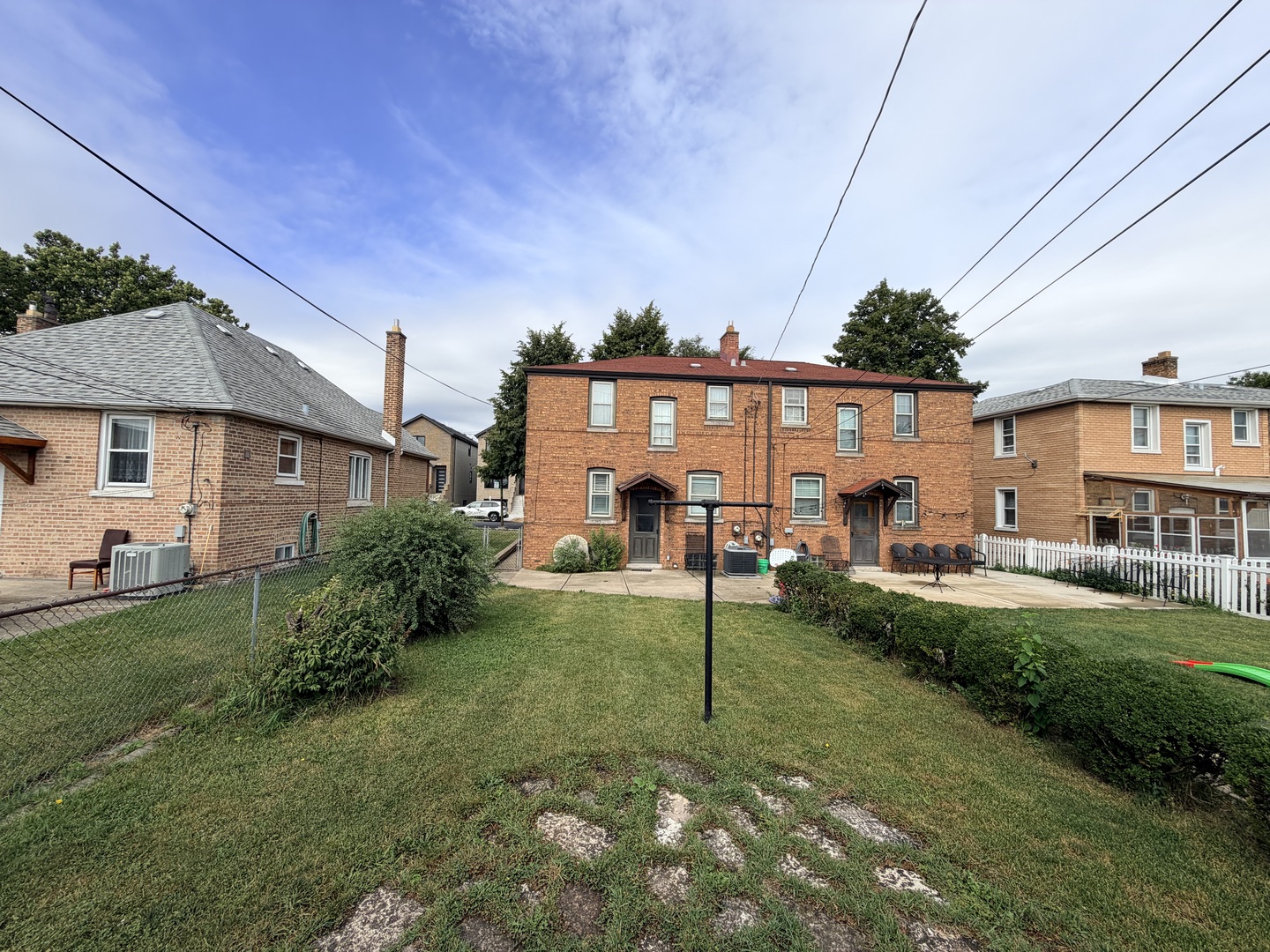 Brick multi-unit building with two rear entrances, patio seating, and a fenced lawn.