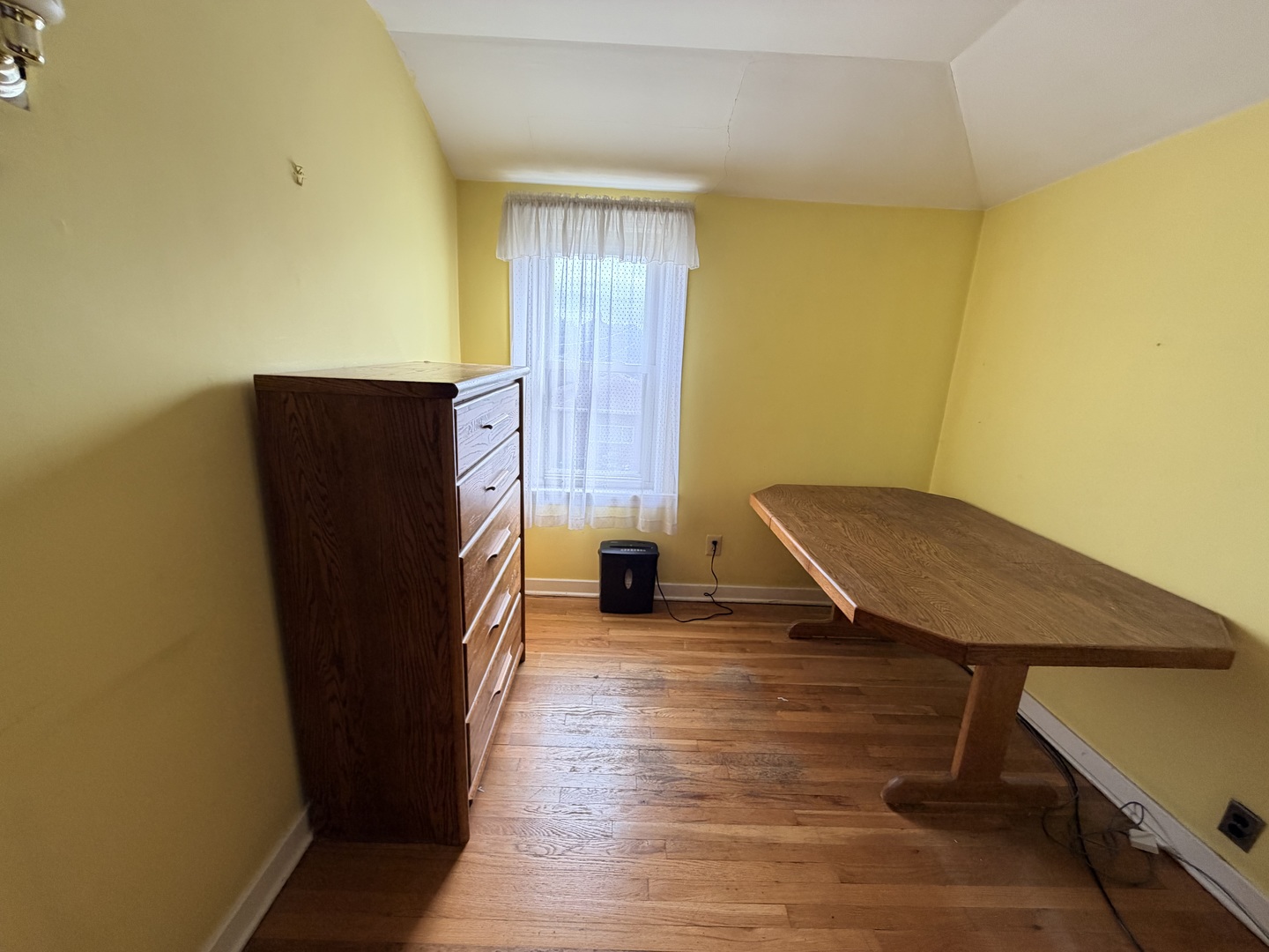 Yellow room with hardwood floors, a wooden dresser, and a long table by the window.