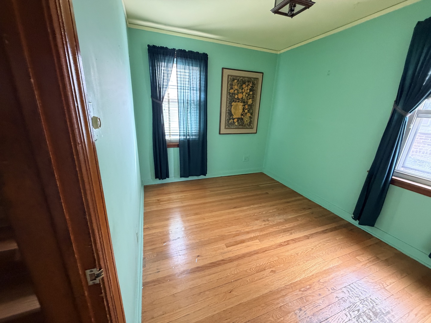 Empty turquoise room with hardwood floors, dark curtains, and two windows.