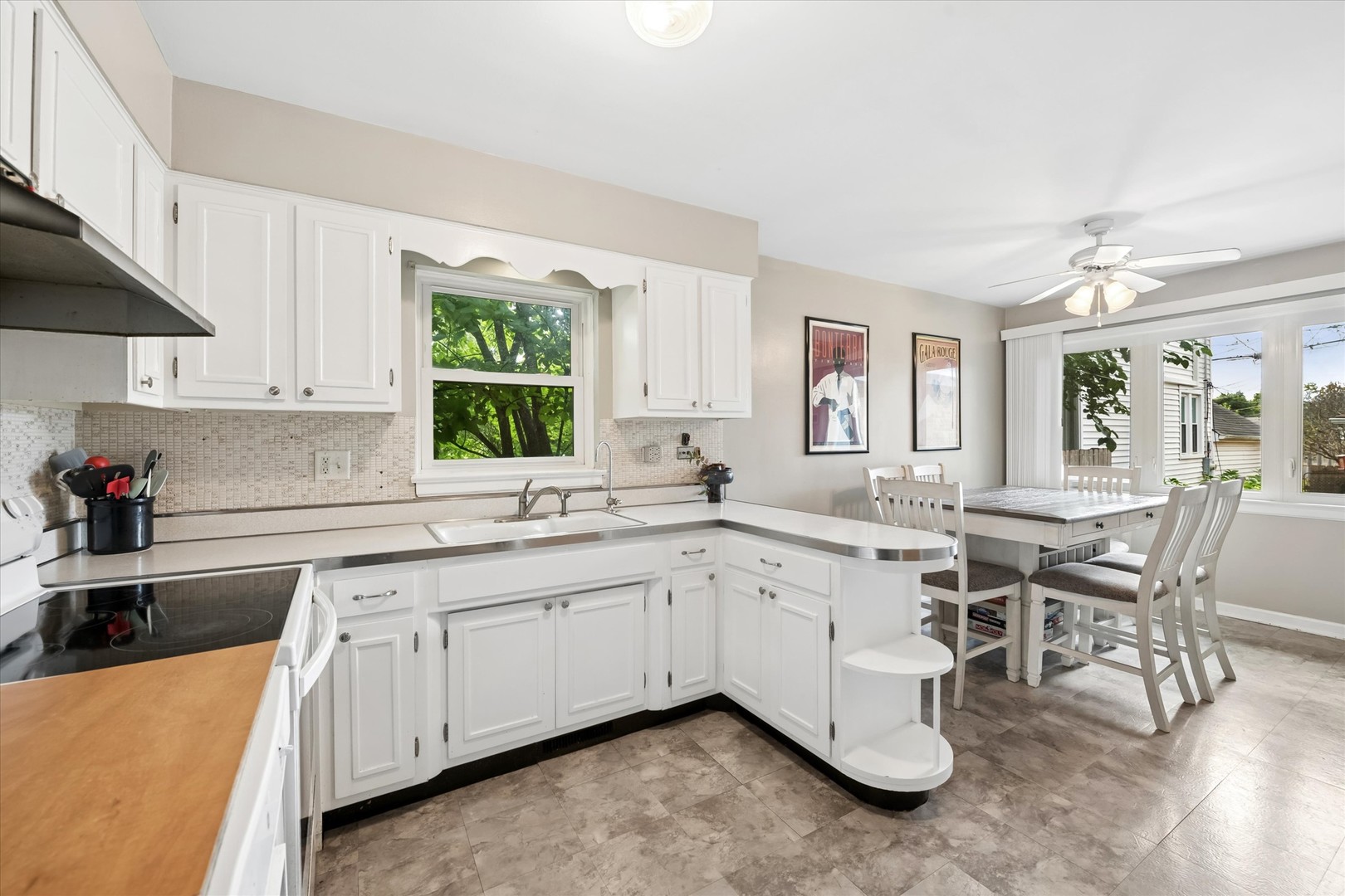 Kitchen with white cabinets and a sink beneath a window, opening to a dining area with large windows.