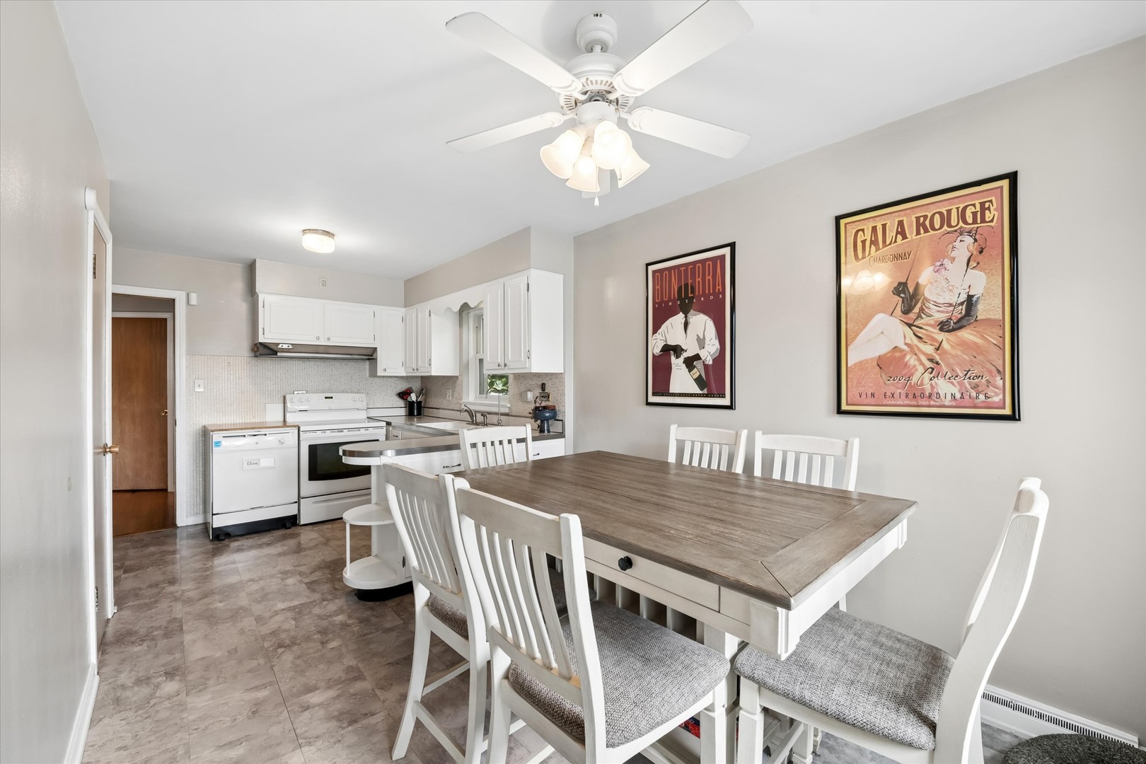 Dining area with a large wood-top table beside a white-cabinet kitchen and two vintage-style wine posters.