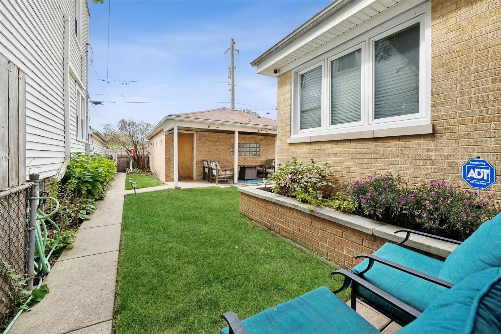 Backyard with a green lawn, flower beds, turquoise lounge chairs, and a covered brick patio.