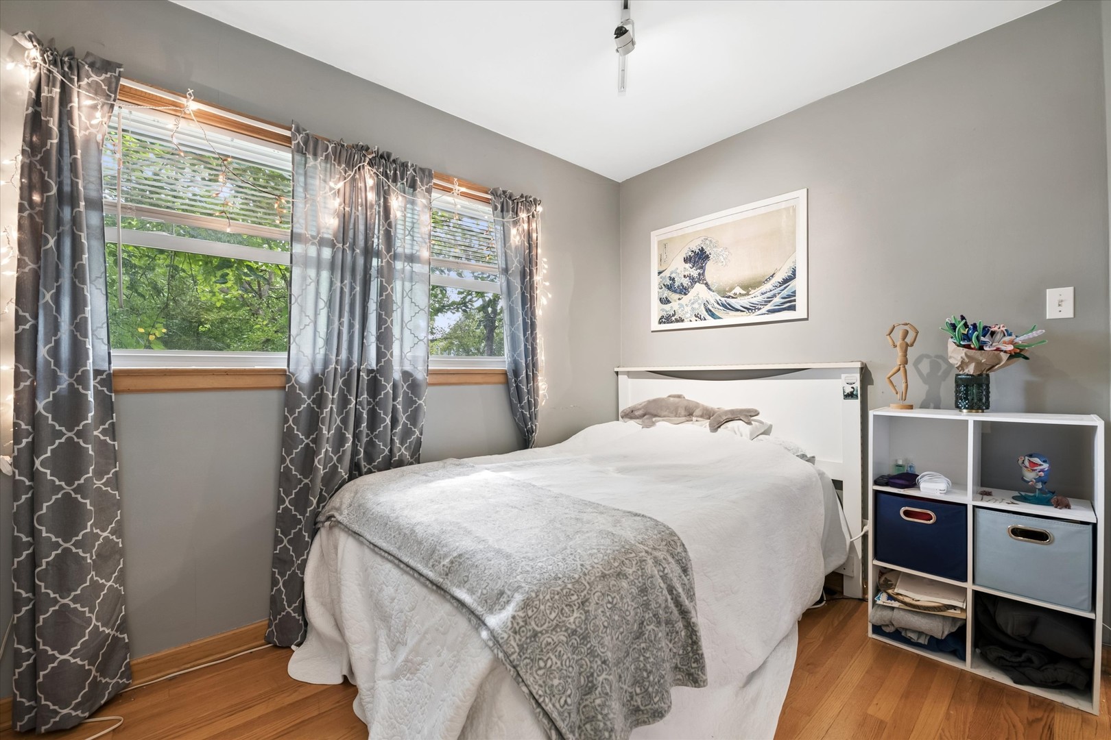Bedroom with a white bed, patterned gray blanket, two curtained windows, and hardwood flooring.