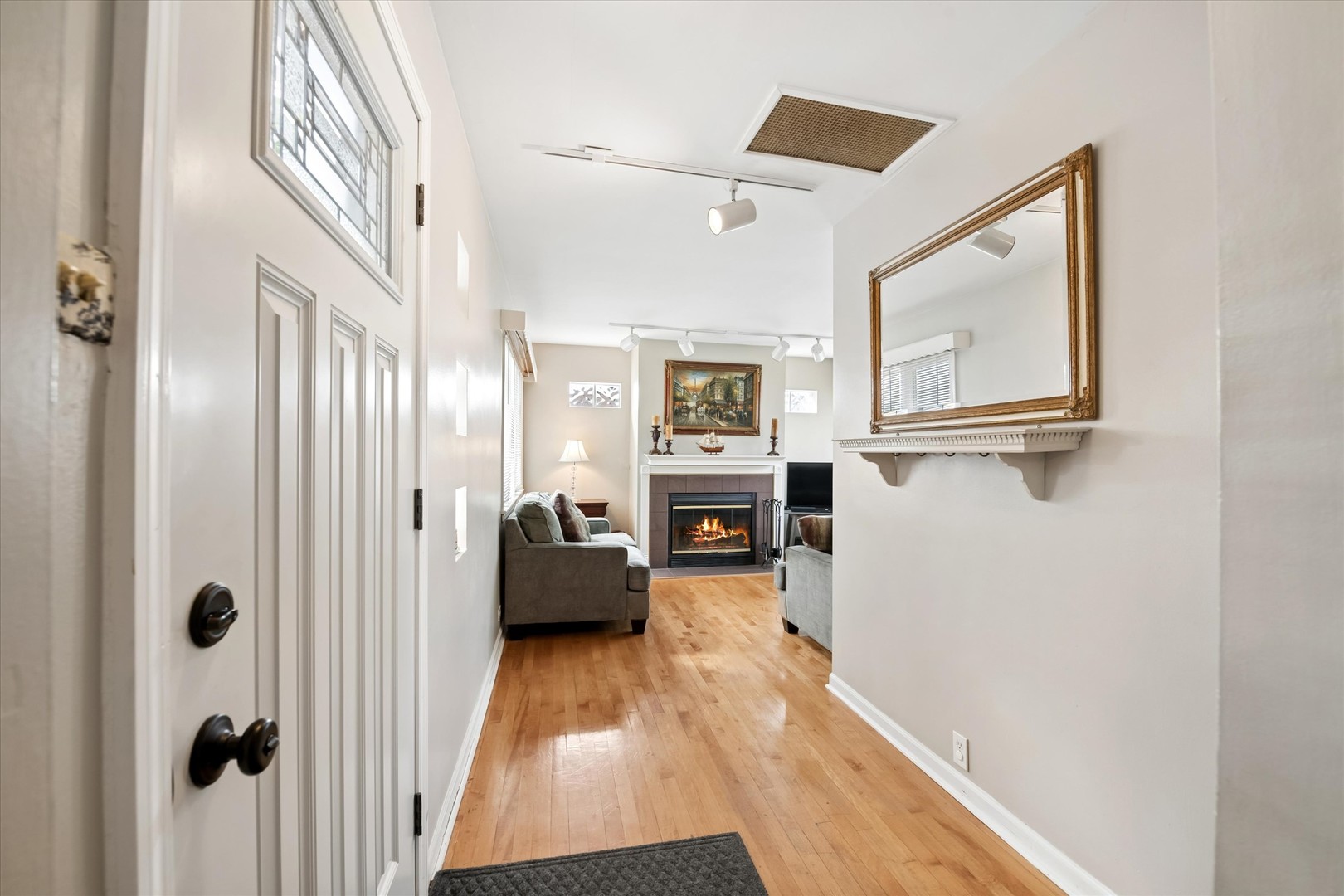 Entryway looking into a living room with hardwood floors, gray sofas, and a lit fireplace.